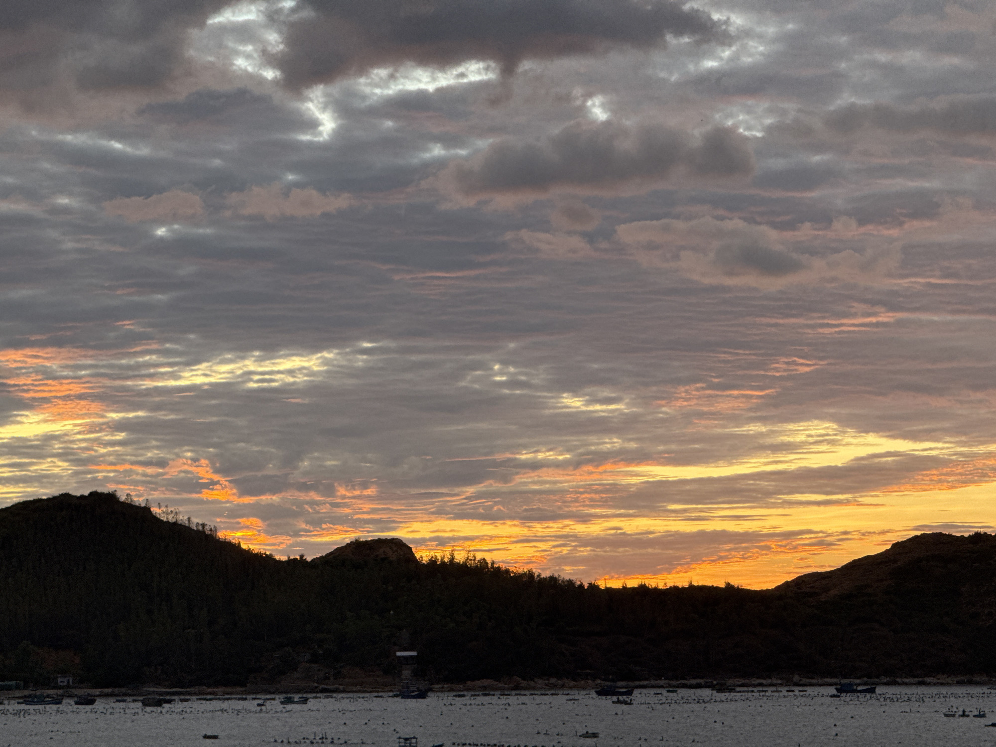 The image shows a sunset over a body of water, with dark, silhouetted hills in the foreground. The sky is filled with dramatic clouds, tinged with orange and yellow from the setting sun, contrasting with the darker grays and purples of the upper sky. Numerous small boats are scattered across the surface of the water, and the hills appear to be covered in dense vegetation. The overall scene is one of natural beauty and tranquility. The location is Unknown, Unknown.