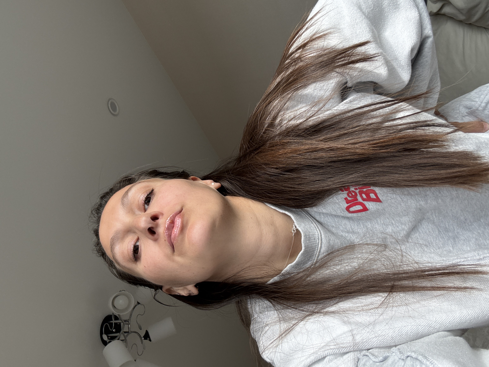 The image is a selfie taken from a low angle. A woman with long, brown hair is the primary subject. She is wearing a light grey sweatshirt with red lettering. Her skin appears fair, and she has glossy lips. She is wearing earrings and a necklace.
The background is a light grey wall with a circular recessed light fixture. In the lower left corner, a decorative wall lamp with multiple frosted glass shades is visible. White bedsheets are partially visible in the top-right corner.
There is no visible evidence suggesting a location in Chaikovsky, Russia. The setting appears to be indoors, likely a bedroom or living space.