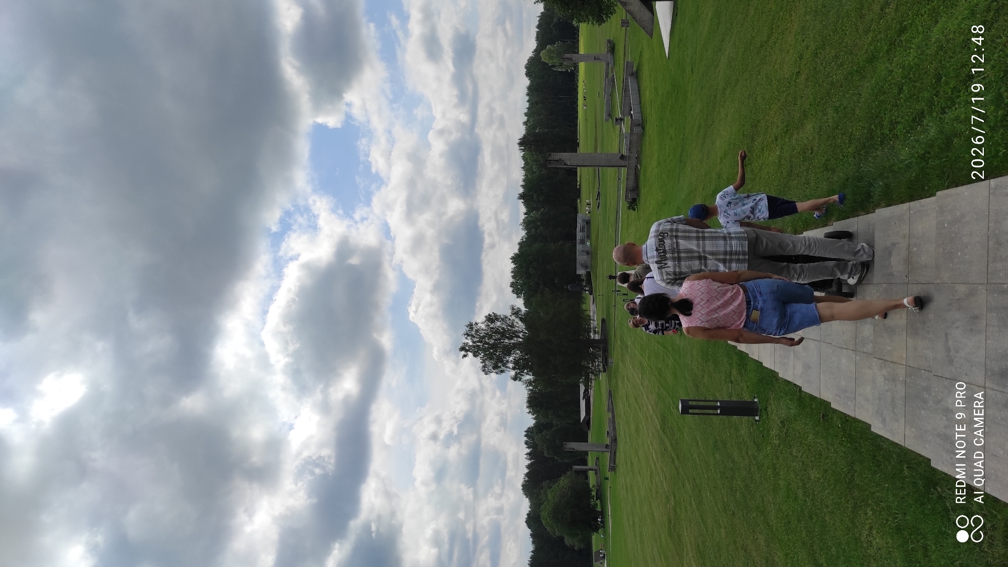 The image shows a group of people walking on a paved path in a park-like setting. The path leads away from the viewer towards a grassy field dotted with various concrete structures and trees. In the foreground, a woman in a pink and white patterned top and denim shorts walks ahead of a man in a plaid shirt and grey pants, who is pushing a stroller. Behind them, a young boy in a blue cap and a patterned t-shirt walks to the right of the path. Several other individuals are visible further down the path, walking towards the background. The sky is filled with dramatic, grey and white clouds, with patches of blue sky visible. The overall environment suggests an outdoor memorial or historical site, possibly related to the ruins or remnants of buildings. The timestamp in the bottom right corner indicates the image was taken on July 19, 2026, at 12:48. The text in the bottom left corner identifies the phone model as "REDMI NOTE 9 PRO AI QUAD CAMERA". This location is identified as being in the vicinity of Каменскі сельскі Савет, Belarus.