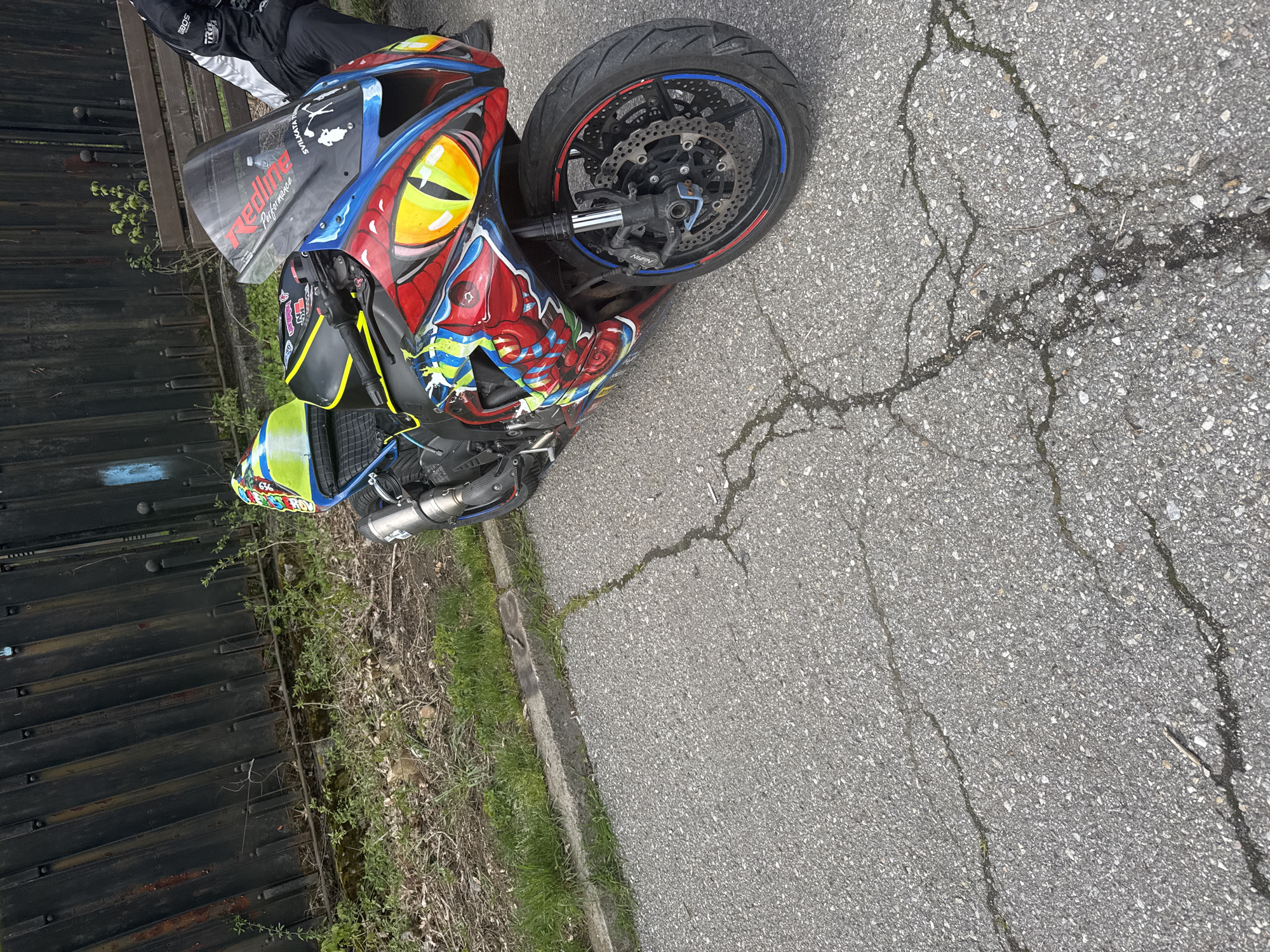A motorcycle is parked on a cracked asphalt surface. The motorcycle is brightly painted with cartoonish depictions of faces and eyes. The front wheel is visible, and the bike appears to be parked perpendicular to the curb. A person wearing protective riding gear stands near the front of the bike. A black metal fence runs along the left side of the frame, with vegetation growing at its base. The location cannot be determined to be Sofia, Bulgaria from the image.