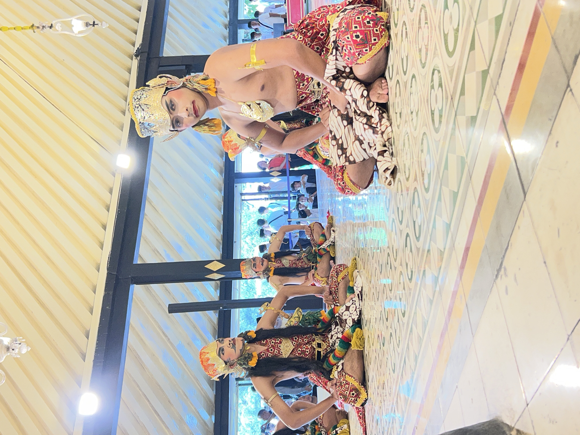 The image shows three individuals dressed in traditional Indonesian dance costumes, kneeling on a patterned tiled floor. The dancers are adorned with elaborate headwear, face paint, and ornate clothing in red, gold, and black. The dancer in the foreground is facing the camera, while the other two are positioned to the left, slightly further back. They appear to be performing a traditional dance, likely in a cultural setting. The background features architectural elements, including a beamed ceiling, dark pillars, and large windows or open doorways revealing blurred figures of an audience. The scene suggests a performance or cultural event taking place indoors, possibly a pavilion or a traditional hall. Based on the traditional attire and the cultural context implied, the location is very likely in Yogyakarta, Indonesia, a city renowned for its rich Javanese culture and traditional arts.