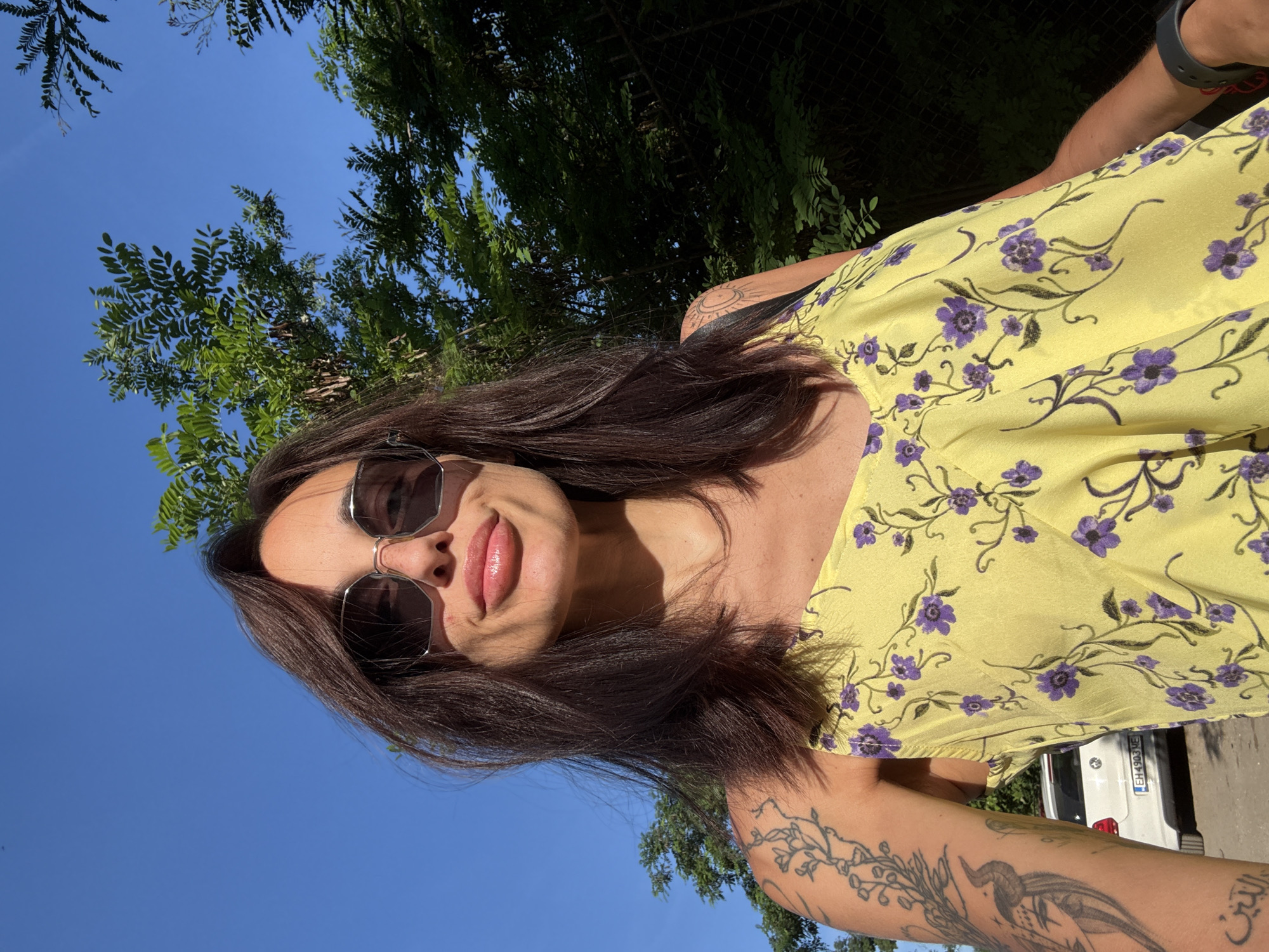A woman with long, dark brown hair and sunglasses is taking a selfie outdoors. She is wearing a yellow dress with a purple floral pattern. On her left arm, she has tattoos. In the background, there are green trees and a blue sky. A white car with a license plate is visible behind her. The image is taken from a low angle, looking up at the woman. The environment appears to be a sunny day, possibly in a park or a street in the city of Pleven, Bulgaria.
