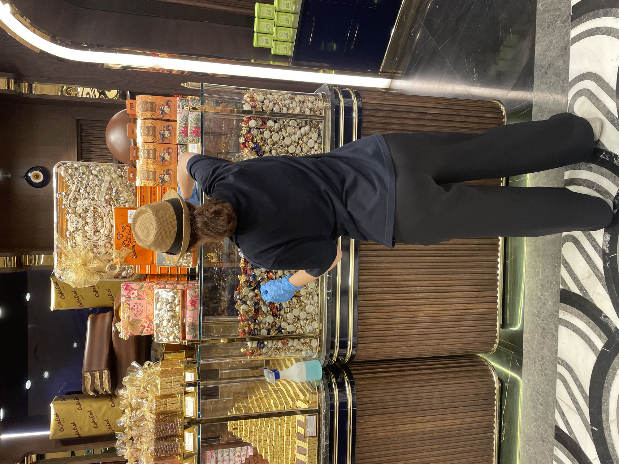 A person wearing a dark blue shirt, dark pants, a straw hat, and blue gloves stands in a well-lit candy shop. The person is holding a spray bottle with blue liquid in their left hand and appears to be spraying the contents of a glass display case filled with individually wrapped candies. The display case is large and cylindrical, with a wooden base. To the left of this display, there are other glass cases filled with golden-colored candies arranged in a pyramid shape, and further left, large boxes of chocolates. The shop's interior features dark wood paneling, gold accents, and a black marble floor with a decorative white and grey pattern. On the right side of the image, there are shelves with several green boxes of candy, and a dark blue cabinet. The overall impression is one of a clean and well-organized confectionery store, possibly located in Kadıköy, Turkey, given the presence of what appear to be Turkish sweets or brands.