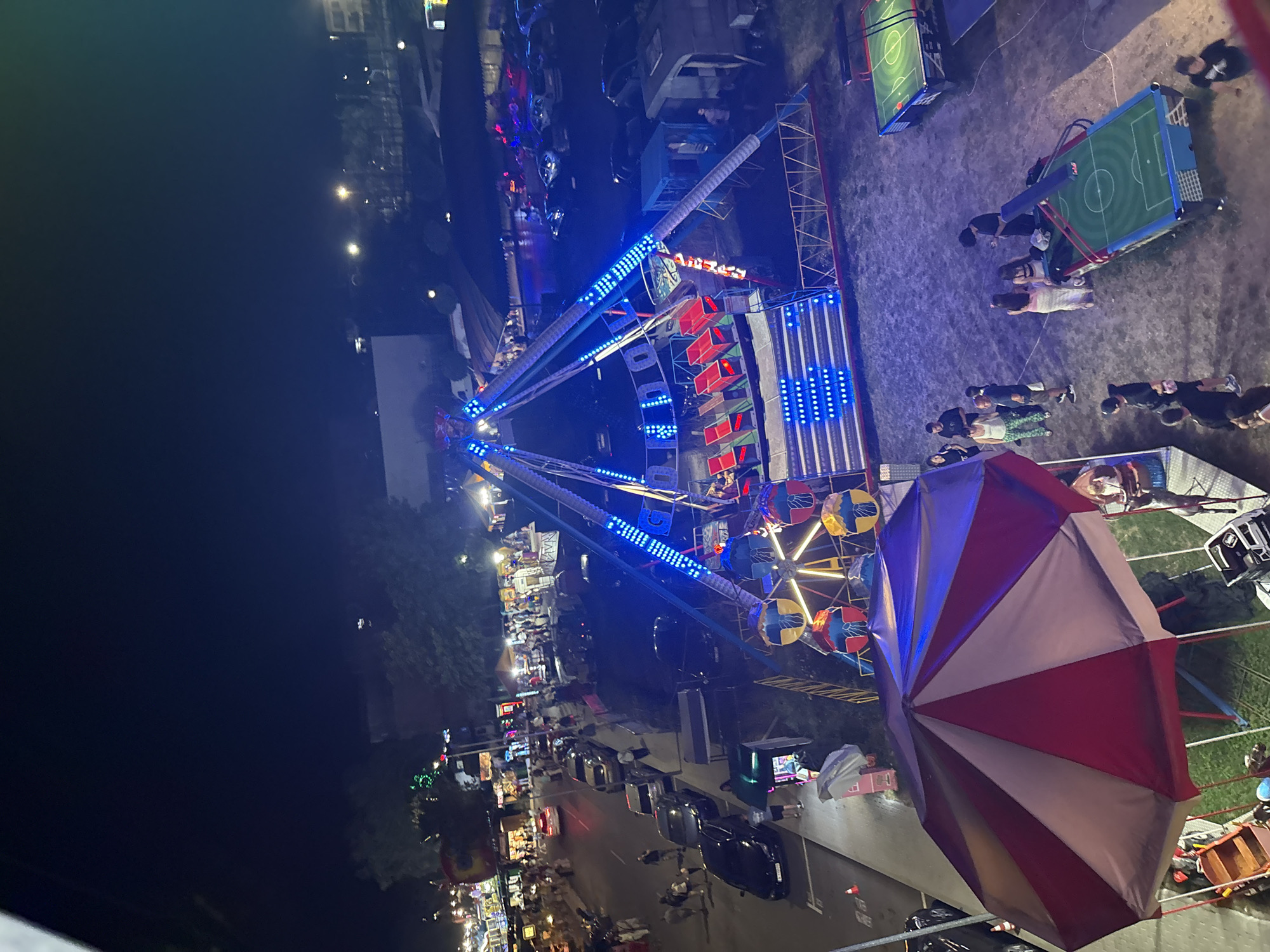 The image depicts an aerial view of an amusement park at night in Primorsko, Bulgaria. A large, pendulum-style ride with blue LED lights is prominent in the center. To the left, a carousel canopy with red and white panels is visible, with a small boat-like ride beneath it. In the background, a street is lined with parked cars and pedestrians, with various amusement stalls and buildings. Several people are seen milling around the park, some near the rides, others near smaller game stalls. Two foosball tables are situated in the foreground to the right. The overall scene is illuminated by artificial lights from the rides, stalls, and streetlights, creating a vibrant, nocturnal atmosphere.