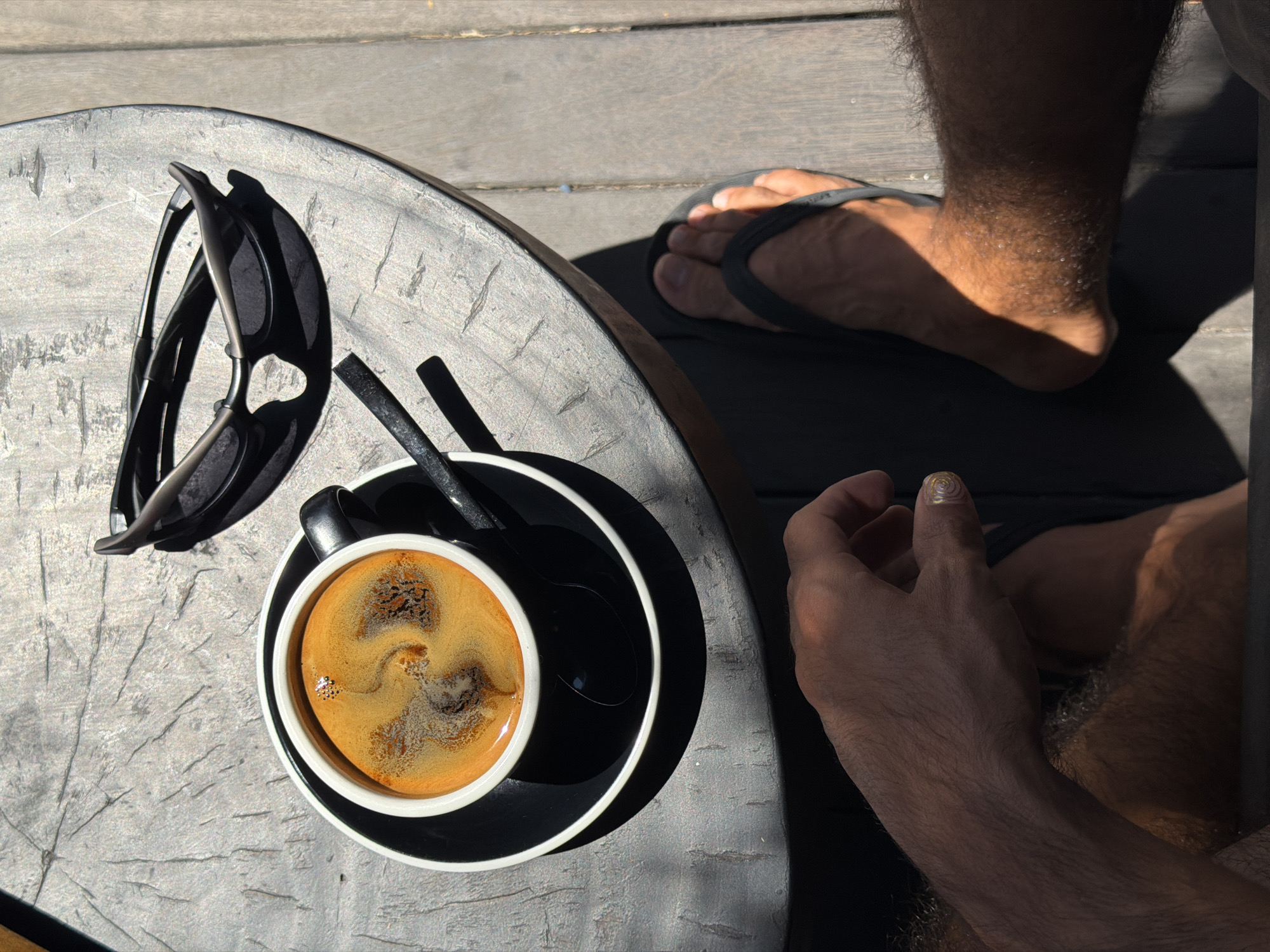 A high-angle, close-up shot shows a black cup of coffee with a foamy crema and a spoon on a black saucer, placed on a rough, textured, circular wooden table. To the right of the coffee cup are a pair of dark sunglasses. In the lower portion of the frame, a person's bare feet in black flip-flops are visible, resting on a wooden deck. The person's right hand, with a gold swirl design on one fingernail, is also visible in the lower left corner. The scene is lit by strong sunlight, casting distinct shadows. The environment appears to be outdoors, possibly at a cafe or restaurant in the city of North Kuta, Indonesia, given the wooden deck and tropical sunlight.