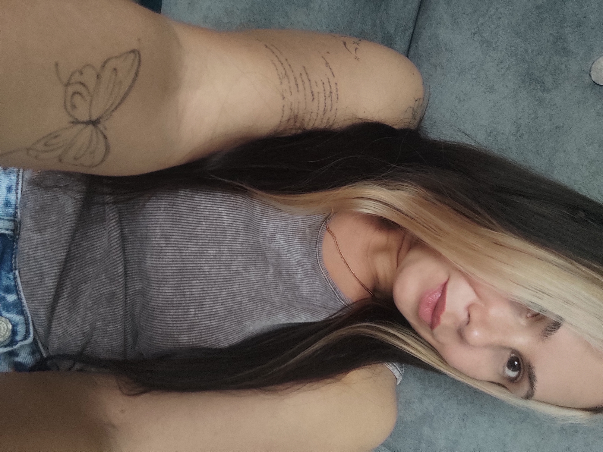 A woman lies on a grey sofa, viewed from above and to the right. Her head is tilted toward the camera. She has dark brown hair with a blonde streak near her face. She wears a grey, striped tank top. The upper part of her left arm, which has two tattoos (a butterfly and script) is visible. The image does not provide sufficient information to place the location within Ольховка, Russia.