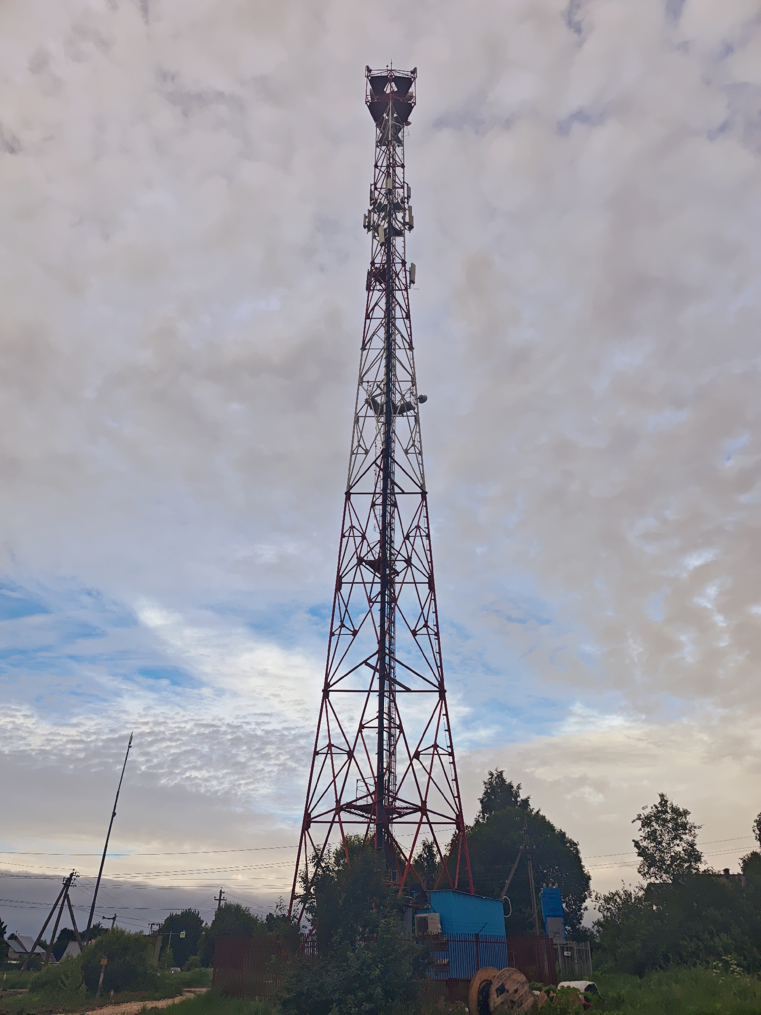 A tall, red and white lattice telecommunications tower dominates the center of the frame. The tower is triangular in cross-section and is covered in various antennas and equipment. It stands against a sky filled with a mix of white, fluffy clouds and patches of blue.
In the foreground and to the right of the tower, there is dense green foliage and trees. A small, light blue metal shed is situated at the base of the tower, with some industrial spools and equipment nearby. To the left of the tower, a dirt path leads towards the foreground. Further to the left, a second, smaller tower structure is visible, along with some houses and more trees. A pedestrian crossing sign is visible in the distance.
The overall impression is of a rural or semi-rural industrial area, possibly on the outskirts of the city of Fedorovskoe, Russia, given the context. The lighting suggests it is either early morning or late afternoon, with soft, diffused light. There are no people visible in the image.