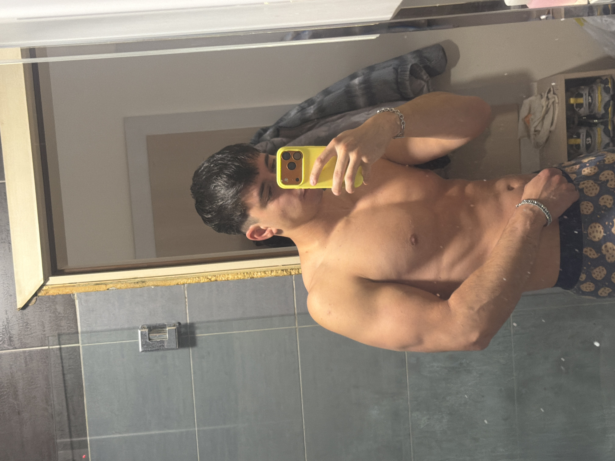 A shirtless male stands in a bathroom, taking a mirror selfie with a yellow-cased phone. He has short, dark hair and is wearing dark underwear patterned with cookie-like designs. A gray and white striped garment is visible in the background. The bathroom features gray wall tiles. There is no indication of the location being Sofia, Bulgaria in the image.