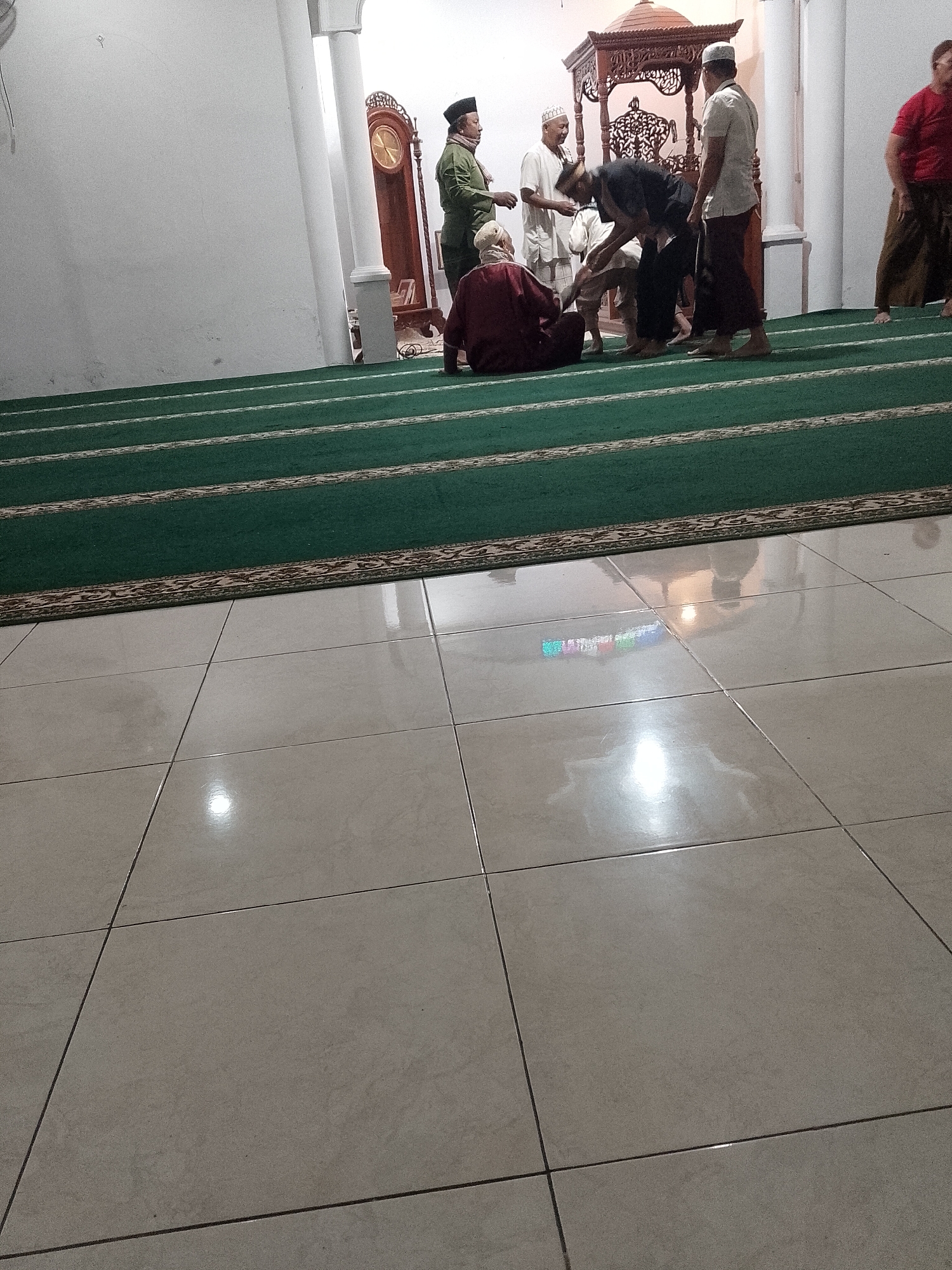 In the city of Ranoyapo, Indonesia, a group of men are gathered in what appears to be a mosque or prayer hall. The floor is covered with light-colored square tiles that reflect the overhead lighting. A vibrant green carpet with decorative borders is laid out on top of the tiled floor, indicating a designated prayer area. Several individuals, dressed in traditional Islamic attire, are present. One person in a maroon garment is seated on the carpet. Others are standing, some engaged in conversation or prayer. In the background, a tall grandfather clock with an ornate wooden casing stands against a white wall. A decorative wooden structure, possibly a pulpit or niche, is also visible. The lighting in the room is bright, casting reflections on the polished tile floor. The scene captures a moment of communal gathering within a religious space.