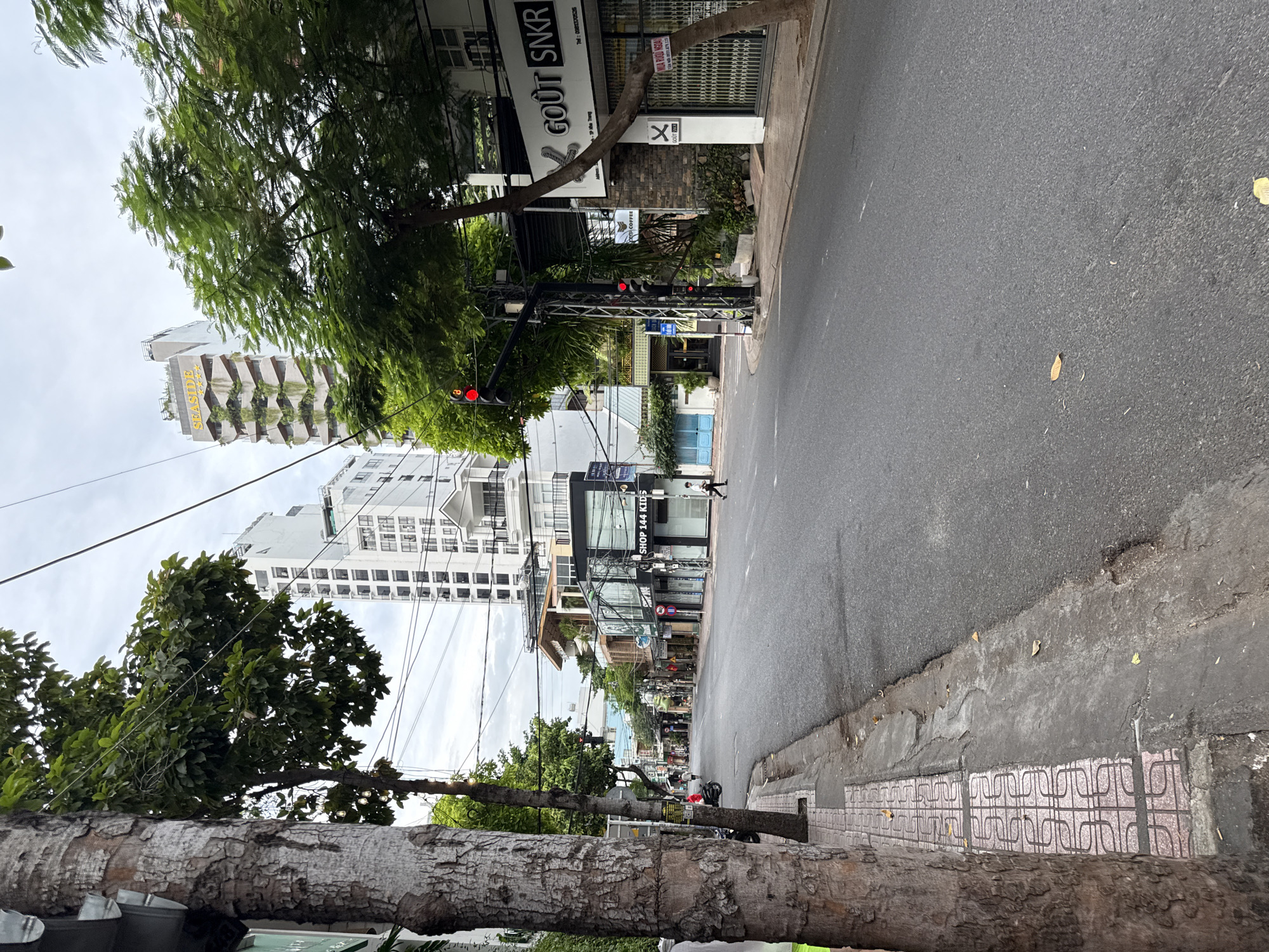 The image captures a street scene in Nha Trang, Vietnam, on an overcast day. The perspective is from street level, looking down a relatively empty road lined with trees and buildings.
On the left, a large tree trunk with rough bark dominates the foreground. Adjacent to the tree is a sidewalk paved with decorative square tiles featuring a repeating pattern of curved lines. Beyond the sidewalk, the road surface is asphalt, showing some wear and tear, with a few fallen leaves scattered about.
The buildings lining the street vary in style and height. On the right side, there are commercial establishments with storefronts. One prominent sign reads "GOÛT SNKR". Another displays "SHOP 144 KIDS". These buildings appear to be multi-story, with some featuring modern architecture and large glass windows. Further down the street, taller buildings, including high-rise structures, are visible in the background, one with a distinctive stepped, green-terraced facade and a sign that says "SEASIDE".
There are a few people visible, but the street is not crowded. A single individual is walking in the middle of the road. Another person is seen on the sidewalk in the distance. A motorbike is parked on the left side of the street. Traffic lights are present at an intersection, with one red light illuminated. Electrical wires crisscross overhead, typical of an urban environment.
The overall impression is one of a quiet urban street during what might be off-peak hours or perhaps due to the weather. The greenery from the trees provides a contrast to the urban infrastructure.