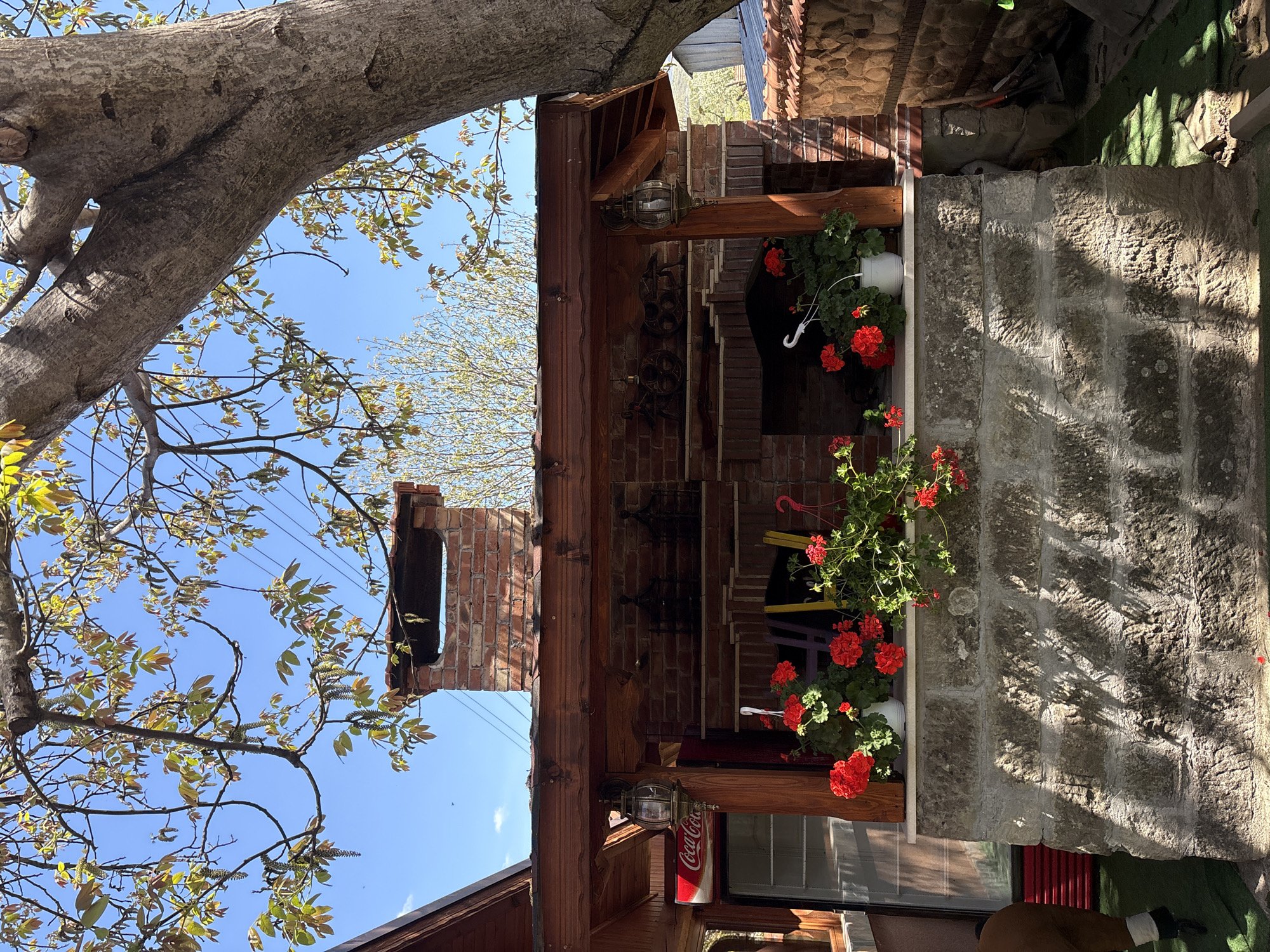The image shows an outdoor structure in what appears to be Kopilovtsi, Bulgaria. The building is constructed of stone and brick, with wooden beams supporting a roof. Hanging flower baskets filled with red geraniums adorn the structure. A brick chimney extends upwards, partially framed by a tree with budding leaves. A vintage Coca-Cola sign is visible below one of the lamps. The setting seems to be a sunny day, as indicated by the bright blue sky visible through the tree branches.