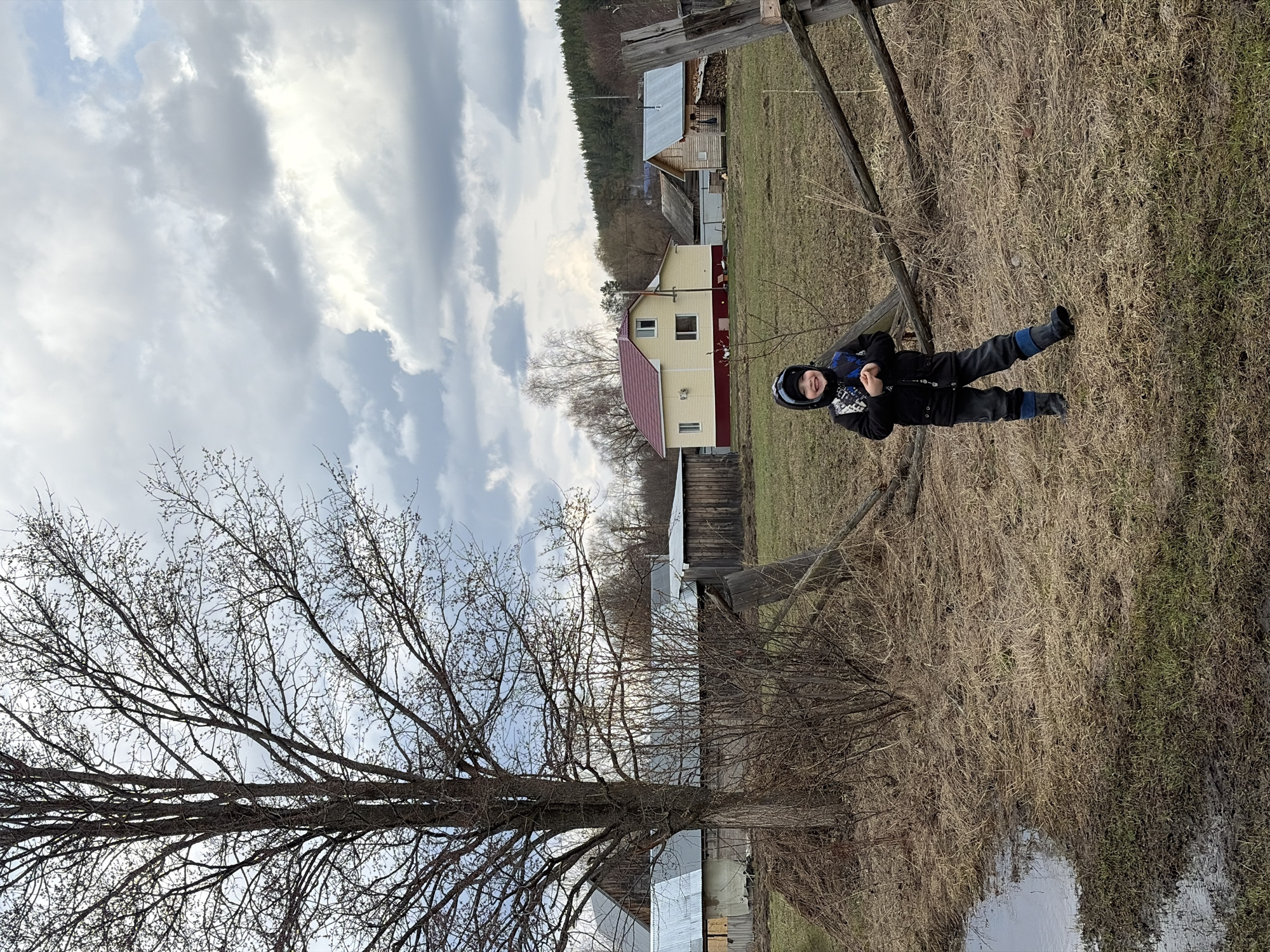 A color photograph, taken outdoors in the city of Кемуль, Russia, shows a young child in the foreground, wearing a dark jacket, dark pants, and a helmet. The child is positioned on a grassy area amidst dry vegetation and is smiling towards the camera.
In the background, a rural landscape is visible. There are several buildings, including a two-story house painted yellow with a red roof. Adjacent to the houses is a forested area on a slight hill. A fence structure runs along the property lines.
The sky is overcast with patches of blue visible. A large, bare tree is prominently featured on the left side of the frame, adding to the overall rustic and somewhat stark aesthetic of the scene. A puddle or body of water reflects the surrounding landscape in the lower right. The photograph appears to be taken during the late fall or early spring, based on the bare trees and dormant vegetation.