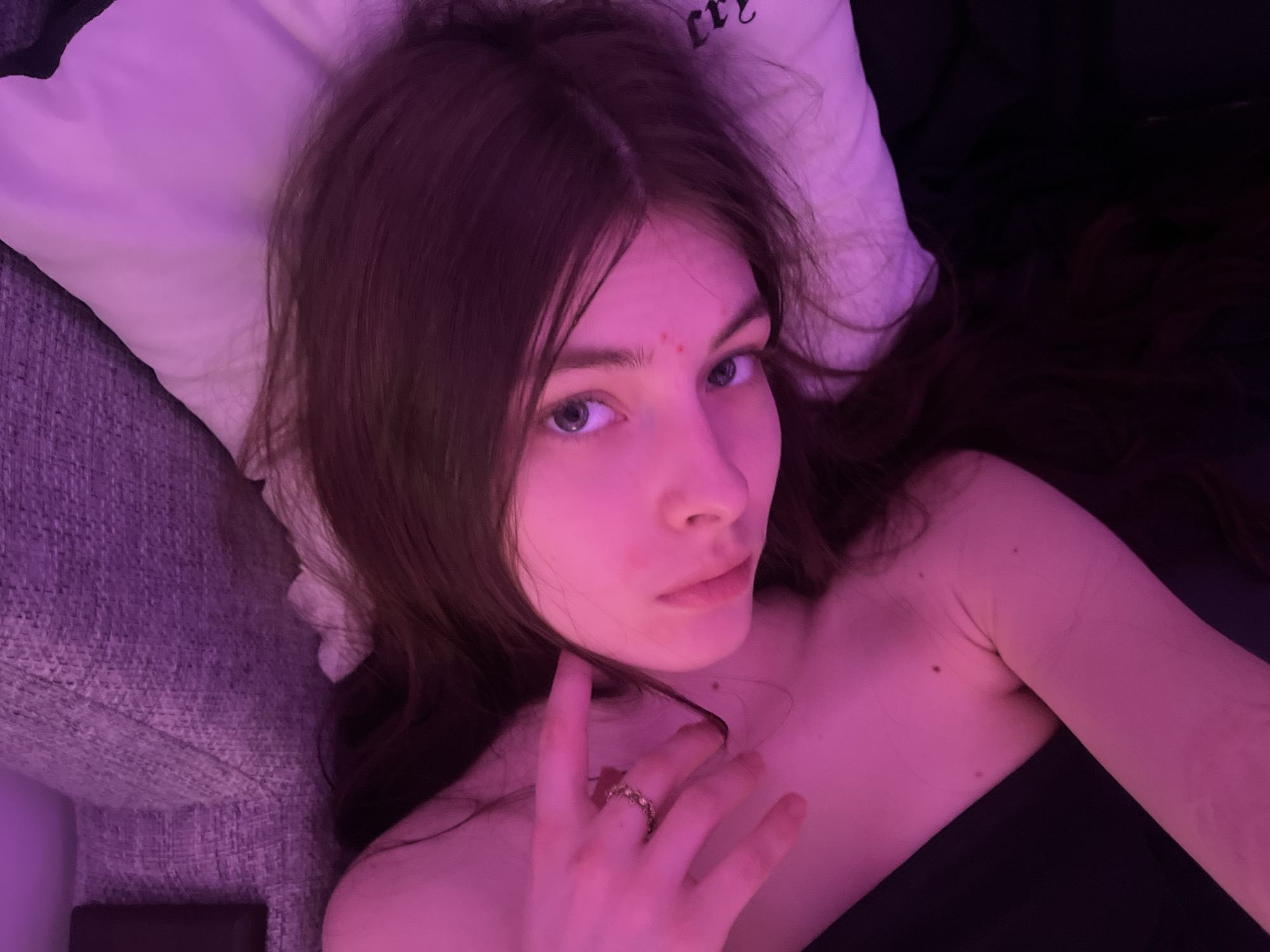 A young woman with long, light brown hair and blue eyes is lying down. She rests on a white pillow, with a gray fabric surface visible nearby. She is wearing a dark-colored top. Her right hand is raised near her neck, with a ring visible on one of her fingers. The lighting is predominantly purple. The location and city are not discernible from the image.