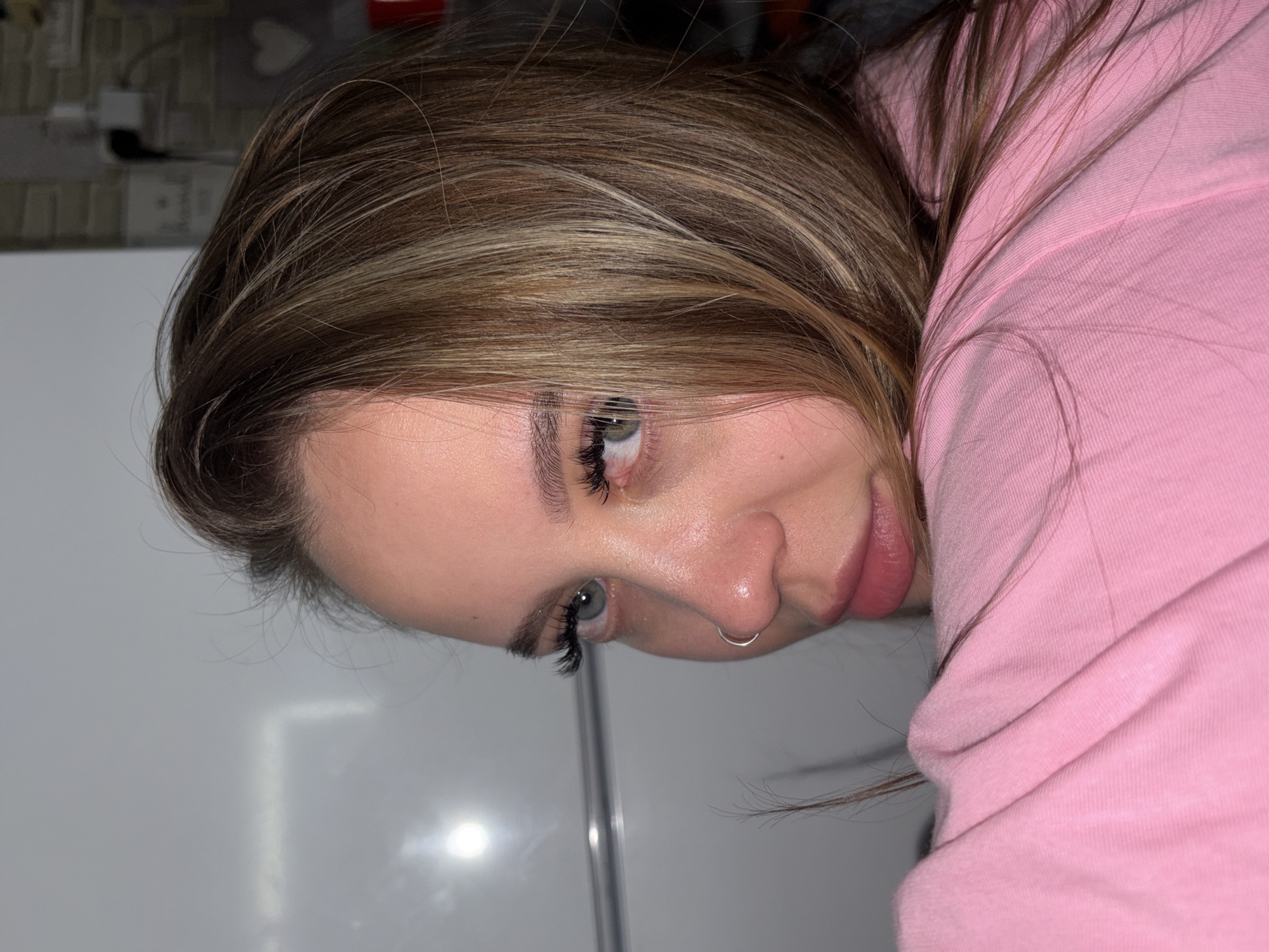 A close-up, angled portrait shows a woman with light brown hair highlighted with blonde streaks. She is wearing a pink hooded sweatshirt. A small, silver hoop is visible in her left nostril. Her eyes appear light colored, possibly green or hazel, with dark eyelashes. A white surface appears in the bottom left corner of the frame, and blurred background elements suggest a kitchen setting with tiled surfaces. The location of the scene is not identifiable as being in the city of Port-Katon, Russia, based on the provided visual information.