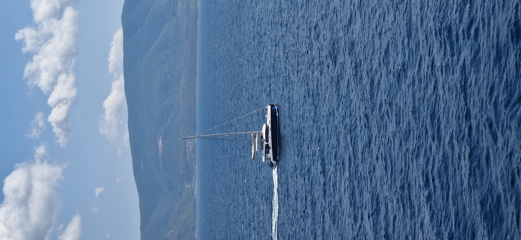 A catamaran sailboat with a dark hull and white superstructure sails on a deep blue sea under a partly cloudy sky. The boat is moving from left to right, leaving a white wake. In the background, a verdant, mountainous coastline stretches across the horizon. The image appears to be taken from a slightly elevated perspective, looking out towards the sea and the distant land. The location is the Municipal Unit of Agioi Theodoroi, Greece, characterized by its coastal mountains and proximity to the sea.