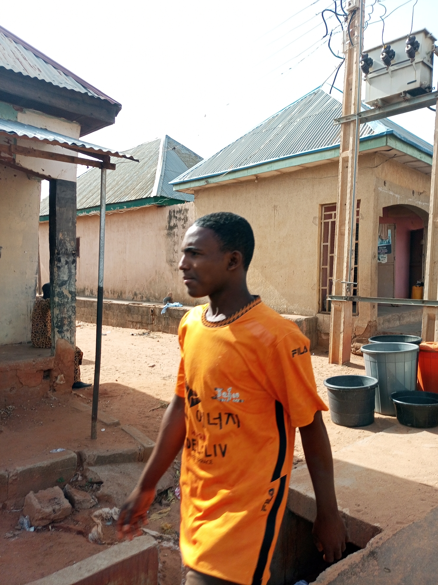 The image shows a man in an orange shirt walking in an outdoor area. The location appears to be a residential area in Tempure, Nigeria.
**People:**
- A man wearing an orange t-shirt with black stripes on the side.
- A person sitting on the porch of a building wearing patterned clothing.
**Environment:**
- Dusty, sandy ground.
- Buildings with corrugated metal roofs.
- A utility pole with a transformer.
**Objects:**
- Several plastic buckets.
- Concrete drains or gutters.
- Electrical wires.
**Actions/Interactions:**
- The man is walking, seemingly in the foreground of the image.
- The other person is sitting still.