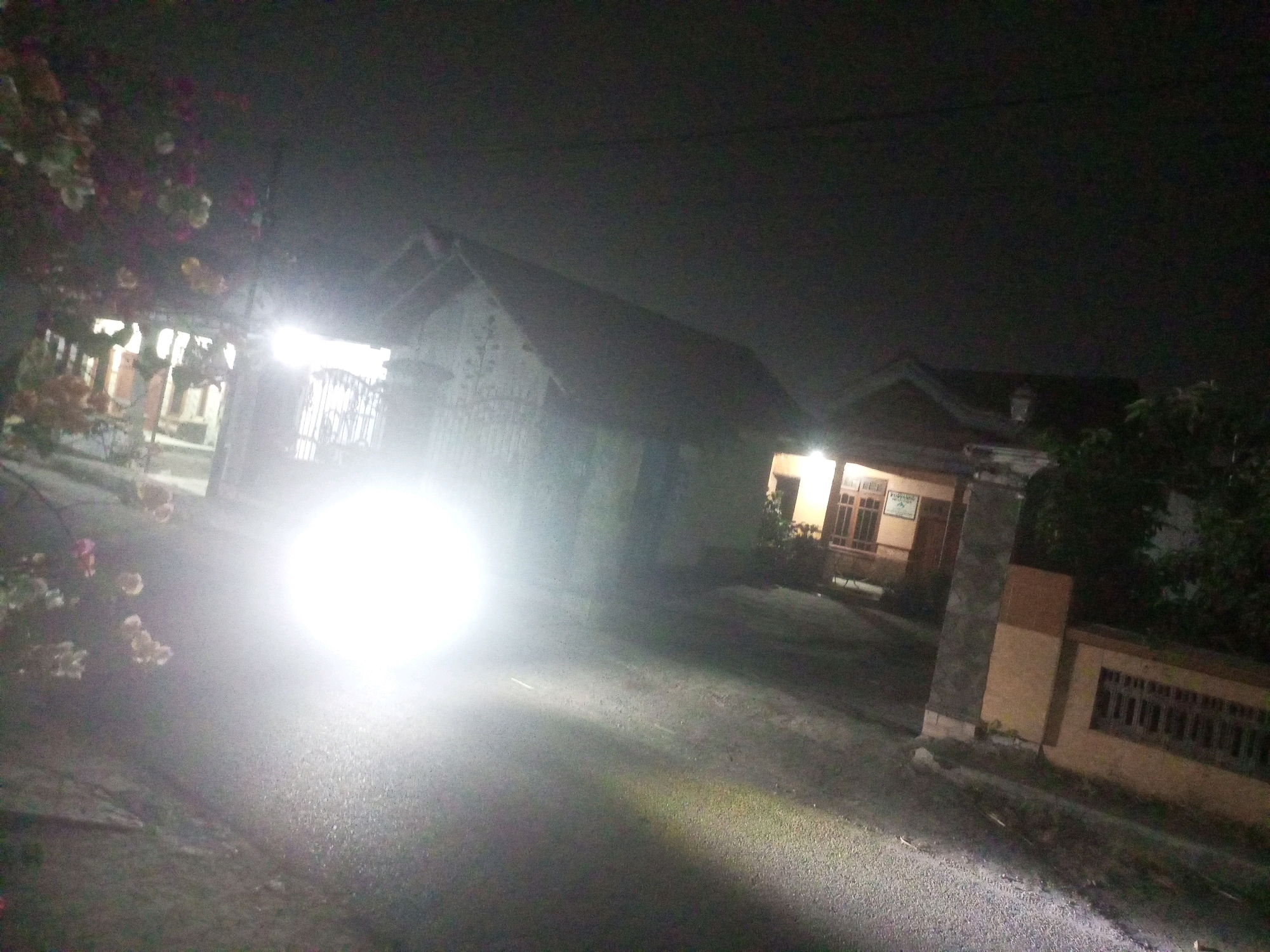 This is a nighttime photograph taken from a street in the city of Ngasem, Indonesia. The image is dominated by a bright, blown-out light source on the left side, likely from a vehicle's headlights or a streetlamp, which illuminates the asphalt road and casts a strong glow. In the background, houses with visible rooflines and windows are dimly lit, suggesting domestic interiors. There are also glimpses of vegetation, including flowering plants on the left. The scene appears to be a residential area at night, with the intense light creating a high-contrast, somewhat obscured view of the surroundings.