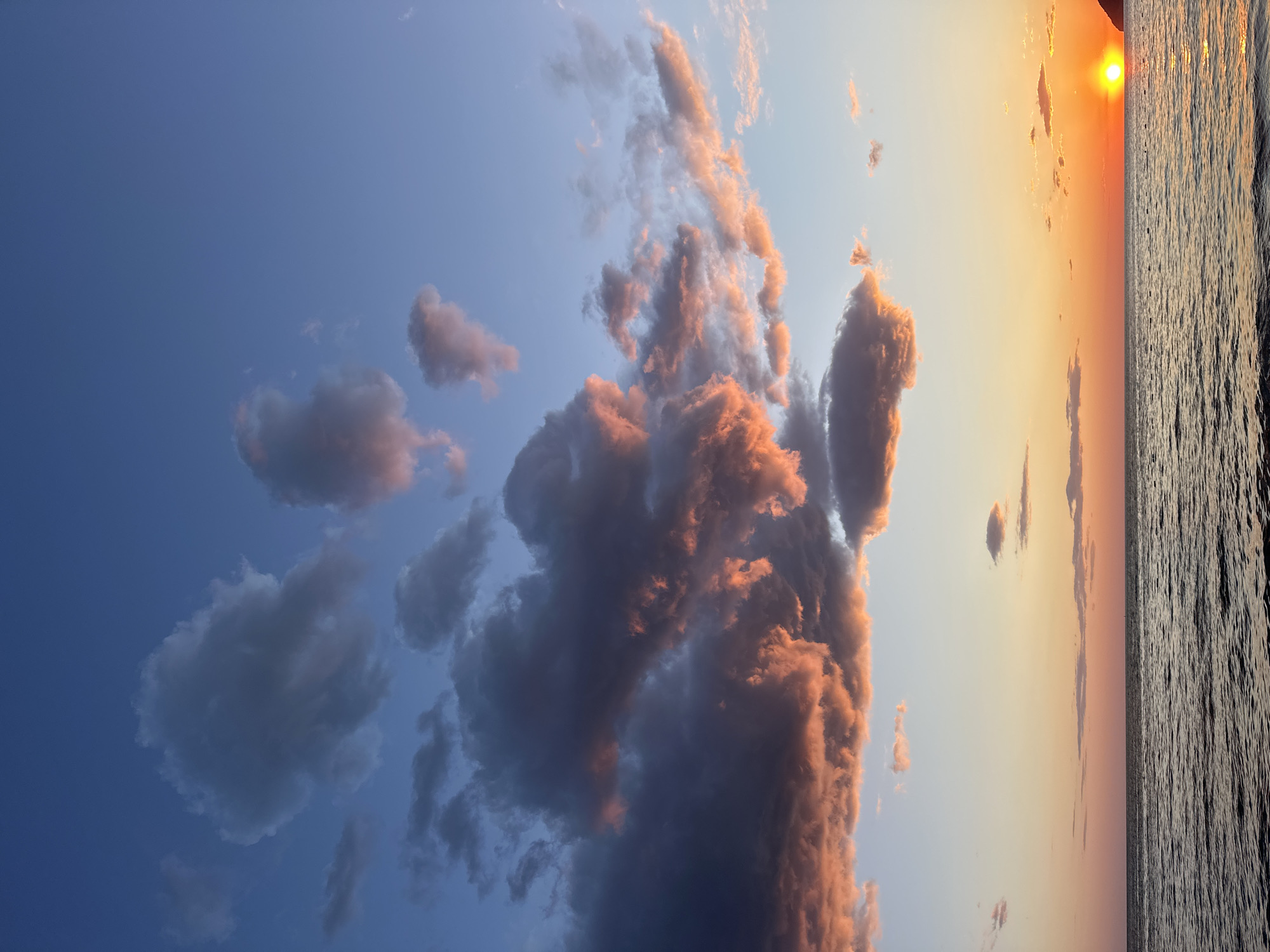 This is a landscape image capturing a vibrant sunset over a body of water, likely the Adriatic Sea near Šušanj, Montenegro.
The upper two-thirds of the image depict a dramatic sky. On the left, the sky is a deep, clear blue, transitioning to a lighter blue and then a warm orange and yellow hue towards the right, closer to the setting sun. A significant formation of cumulus clouds dominates the central sky. These clouds are dramatically lit by the low sun, with their edges and upper parts glowing in warm oranges, pinks, and yellows, while their undersides and shadowed masses appear dark grey and purplish. Smaller, darker clouds are scattered across the deeper blue sky on the left, and wispy clouds catch the golden light closer to the sun on the right.
A bright, intense orange-yellow sun, partially obscured, is visible near the top right corner, casting its golden light across the scene.
The bottom third of the image is occupied by a vast body of water. Its surface is textured with numerous small waves or ripples, which reflect the sun's light in shimmering, golden streaks, creating a luminous effect on the water.
The horizon line is clear and separates the sky from the water, extending horizontally across the image. The scene captures the late afternoon or sunset period, characterized by the warm, intense colors of the sky and the low position of the sun. There is no visible text in the image.