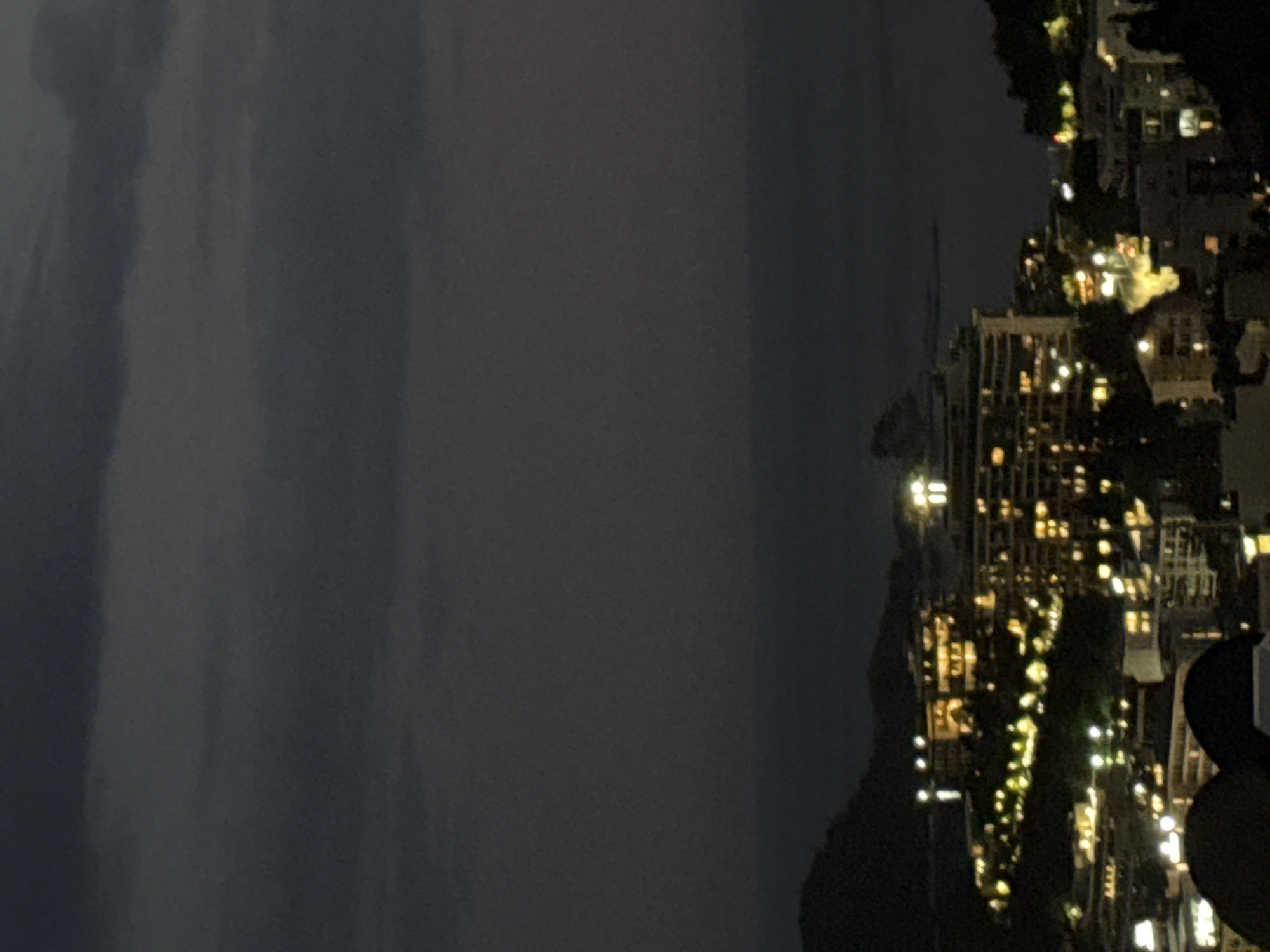 The image depicts a nighttime view of a city illuminated by artificial lights, situated on a hillside overlooking a body of water. The city lights form a dense pattern across the landscape, indicating a populated area. Buildings of various sizes and structures are visible, some appearing to be hotels or residential complexes, with numerous windows lit from within. The sky is dark and overcast, with clouds obscuring any stars or moon. The overall impression is that of an urban environment at night, possibly a resort town given the proximity to the water. Based on the provided information, the location is identified as Bečići, Montenegro.