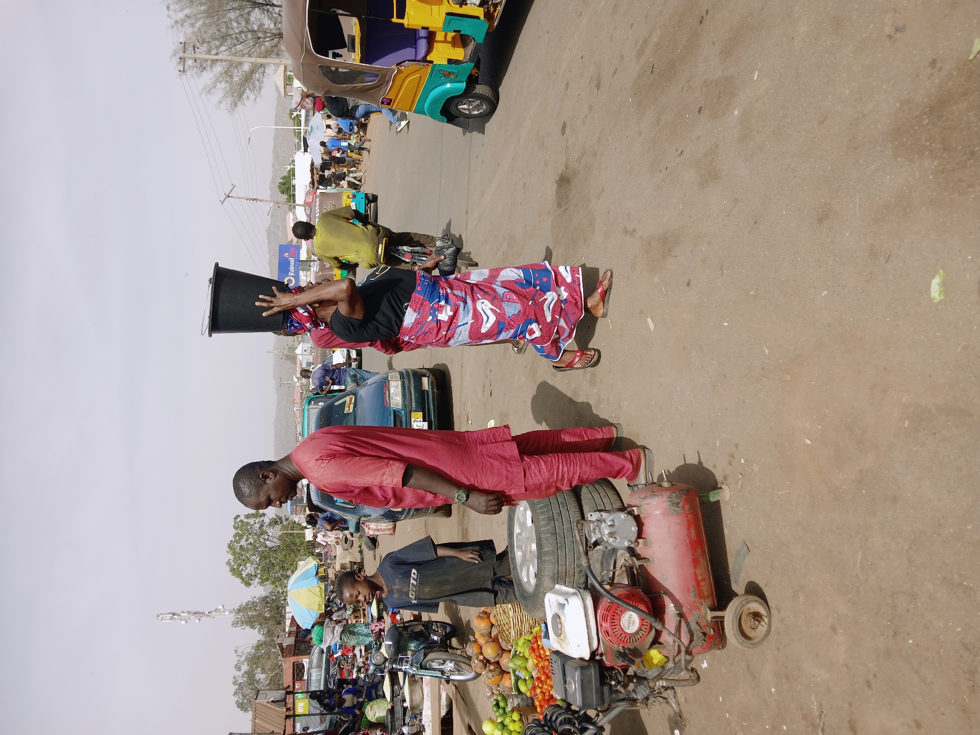 The image shows a street scene in Rafin Par, Nigeria. Two people are prominently featured: one man in a red ensemble standing next to a small, wheeled machine or cart laden with fruits, vegetables, and a spare tire, and another person in a black shirt and red and white patterned wrap carrying a large black tub on their head. A young person in a black shirt stands near the man in red. Various vehicles, including a three-wheeled vehicle, and people populate the background, suggesting a market or busy area. There is a cell tower in the background as well as shops or covered areas with umbrellas, which suggests that they may be selling goods at market.