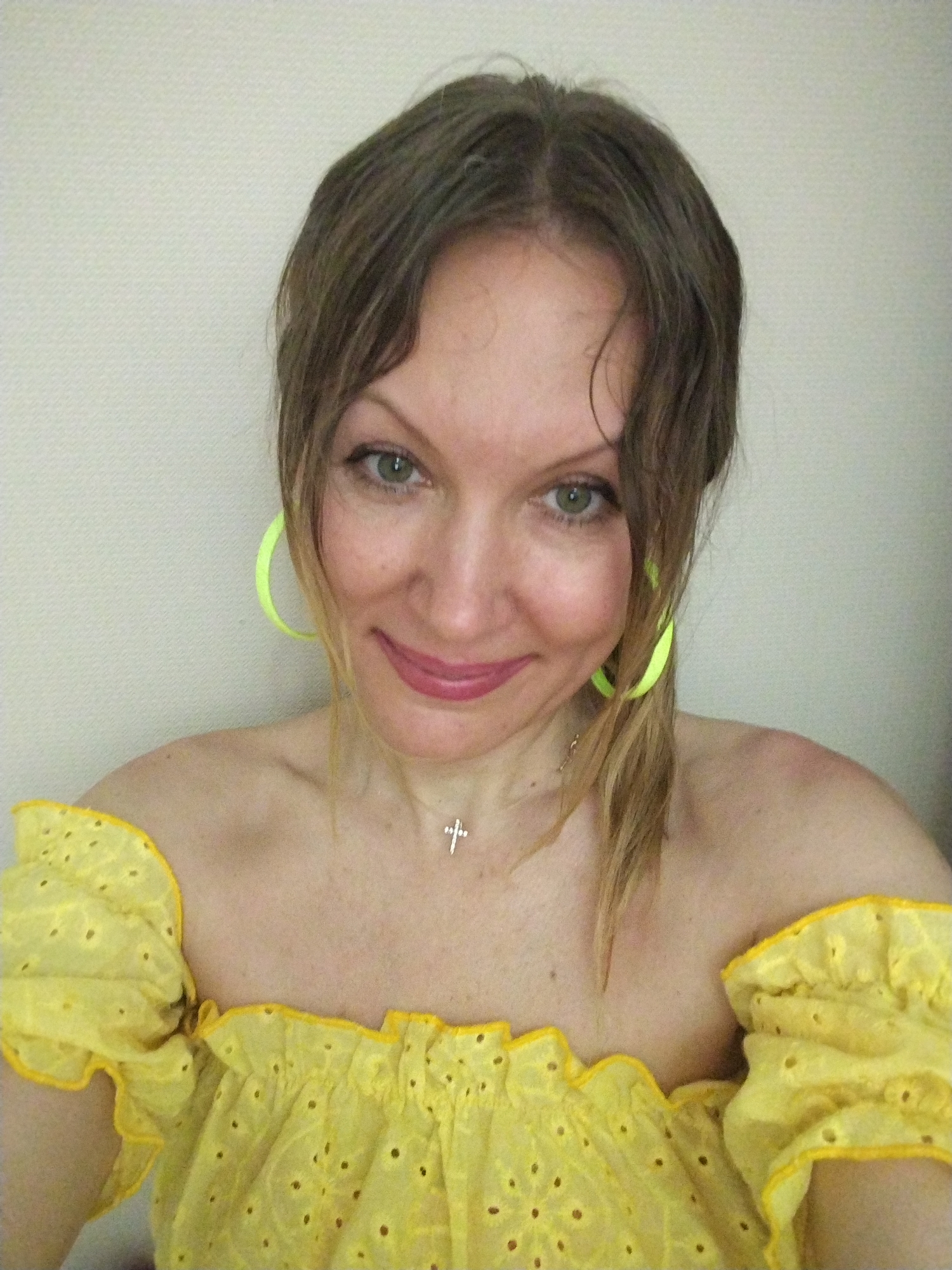 This is a close-up headshot of a woman, presented as a selfie, taken indoors in Krasnodar, Russia. The woman has brown hair, styled loosely with some strands framing her face, and prominent green eyes. She is looking directly at the camera with a slight, pleasant smile, wearing light makeup including pinkish lipstick.
She is dressed in a bright yellow, off-the-shoulder top featuring ruffled edges and eyelet embroidery. A pair of large, circular neon green hoop earrings are visible on both ears, adding a vibrant accent. Around her neck, she wears a delicate silver chain necklace with a small, stylized cross pendant.
The background is a plain, light-colored wall, possibly off-white or very light beige, suggesting an uncluttered indoor environment. The lighting appears natural, consistent with daytime. There is no visible text in the image. The woman's overall expression conveys a calm and contented mood.