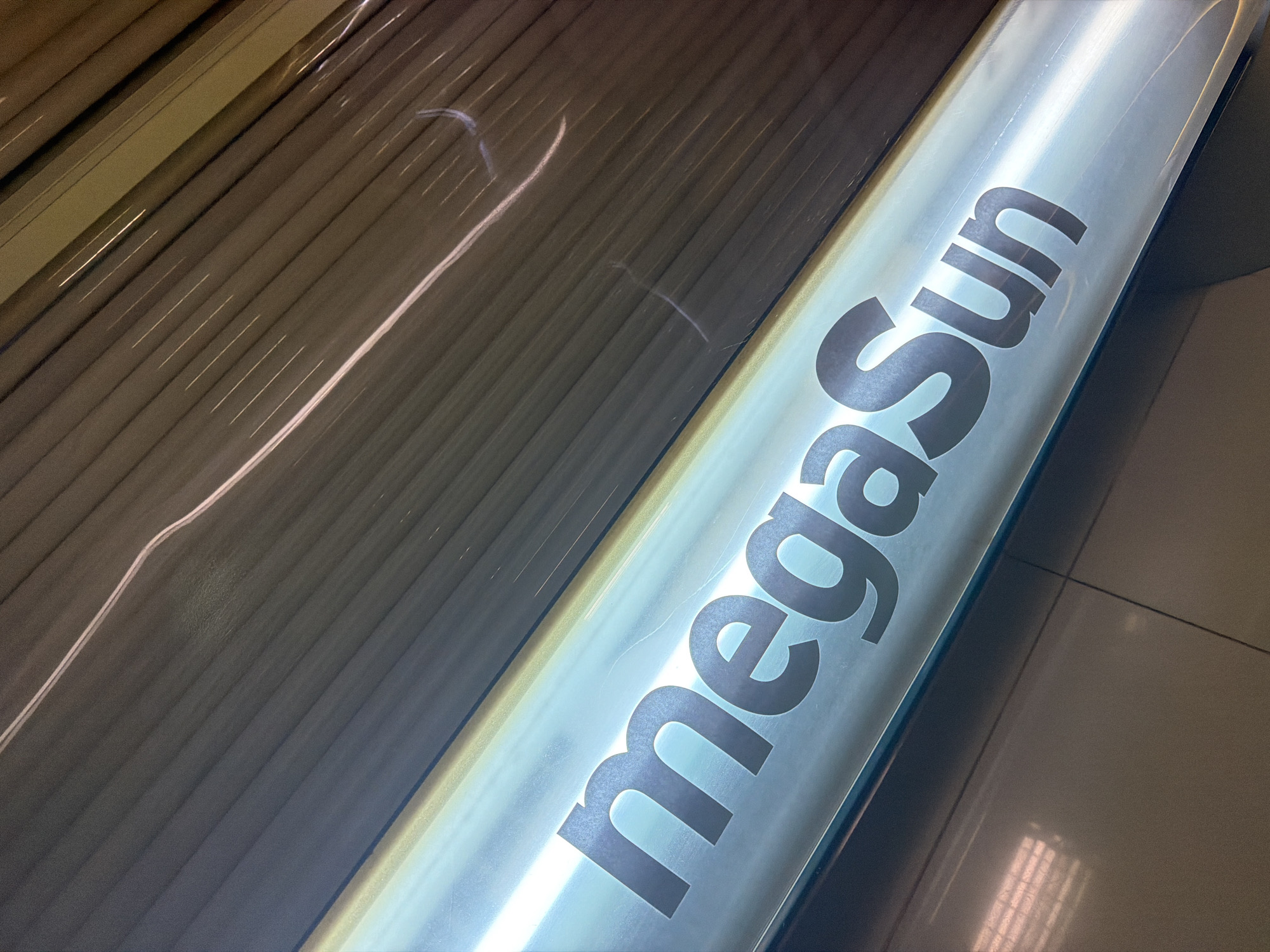 The image shows a close-up, angled view of a tanning bed with the brand name "megaSun" illuminated in large, dark gray letters against a bright, white background. The tanning bed appears to be part of a salon or spa. The background is composed of the ribbed, beige interior of the tanning bed, with some light streaks suggesting movement or reflections. The overall impression is one of modern, clean design. The location is specified as Kyiv, Ukraine.