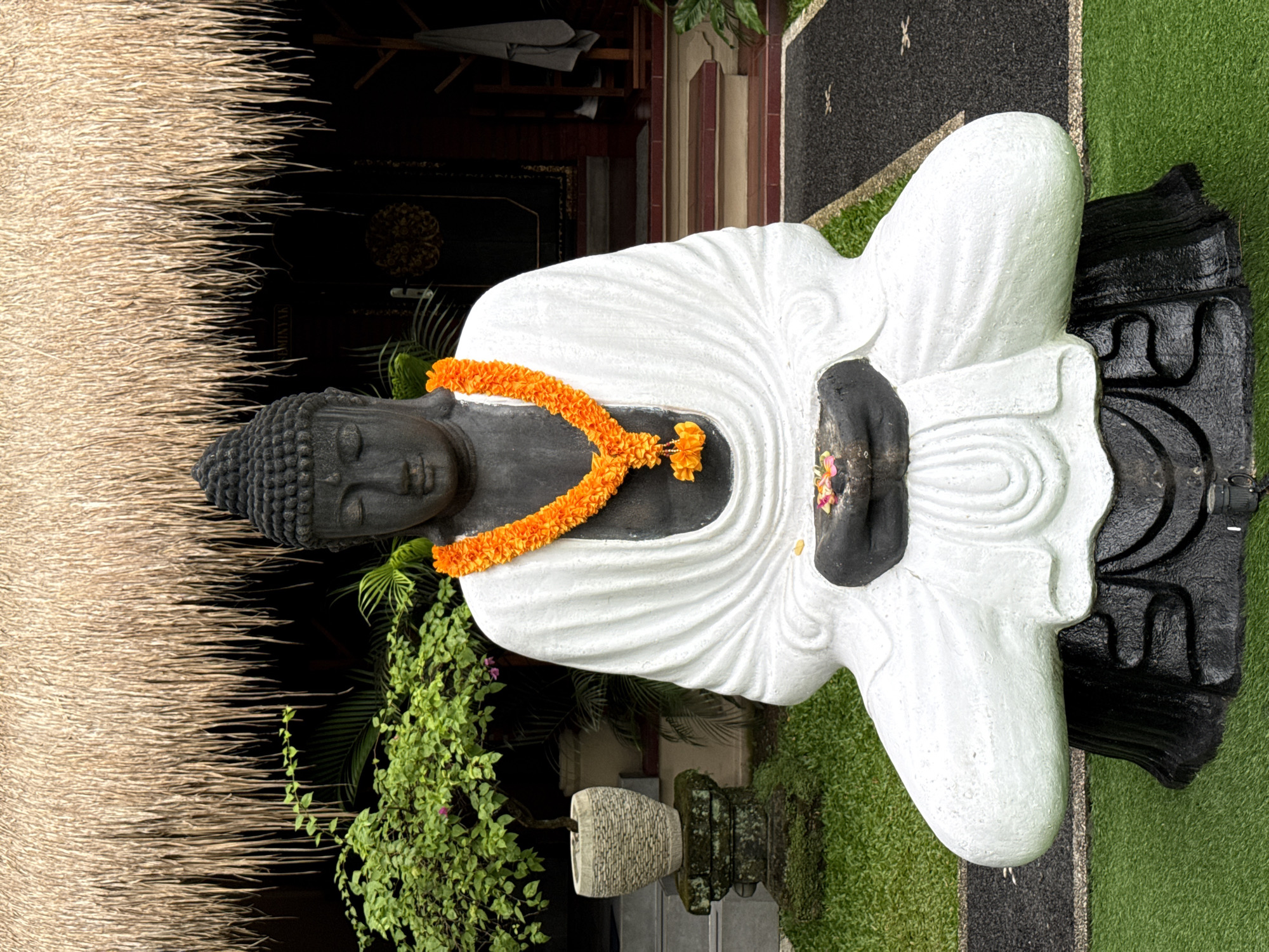 A large stone statue of Buddha is the central subject, depicted in an outdoor setting in Ubud District, Indonesia. The Buddha statue features a dark, textured head and hands, while its body is draped in a flowing white robe. A vibrant orange marigold garland is placed around its neck, and small pink/purple flowers with a green leaf are carefully arranged in its clasped hands, signifying an offering or devotion.
The statue is surrounded by lush greenery, including various plants and climbing vines. Behind the Buddha, a traditional thatched (alang-alang) roof is prominently visible on the left, indicative of Balinese architecture. A dark, intricately carved structure, possibly a wooden door or wall, stands in the background. To the right, the ground is covered with artificial green grass and dark gravel pathways. A decorative stone pot or vessel is visible to the left of the Buddha. The scene is illuminated by natural light, suggesting it is daytime. A faint and partially obscured word, possibly "WARNING," is visible on a dark surface in the background. The overall atmosphere conveys peace and spiritual reverence.