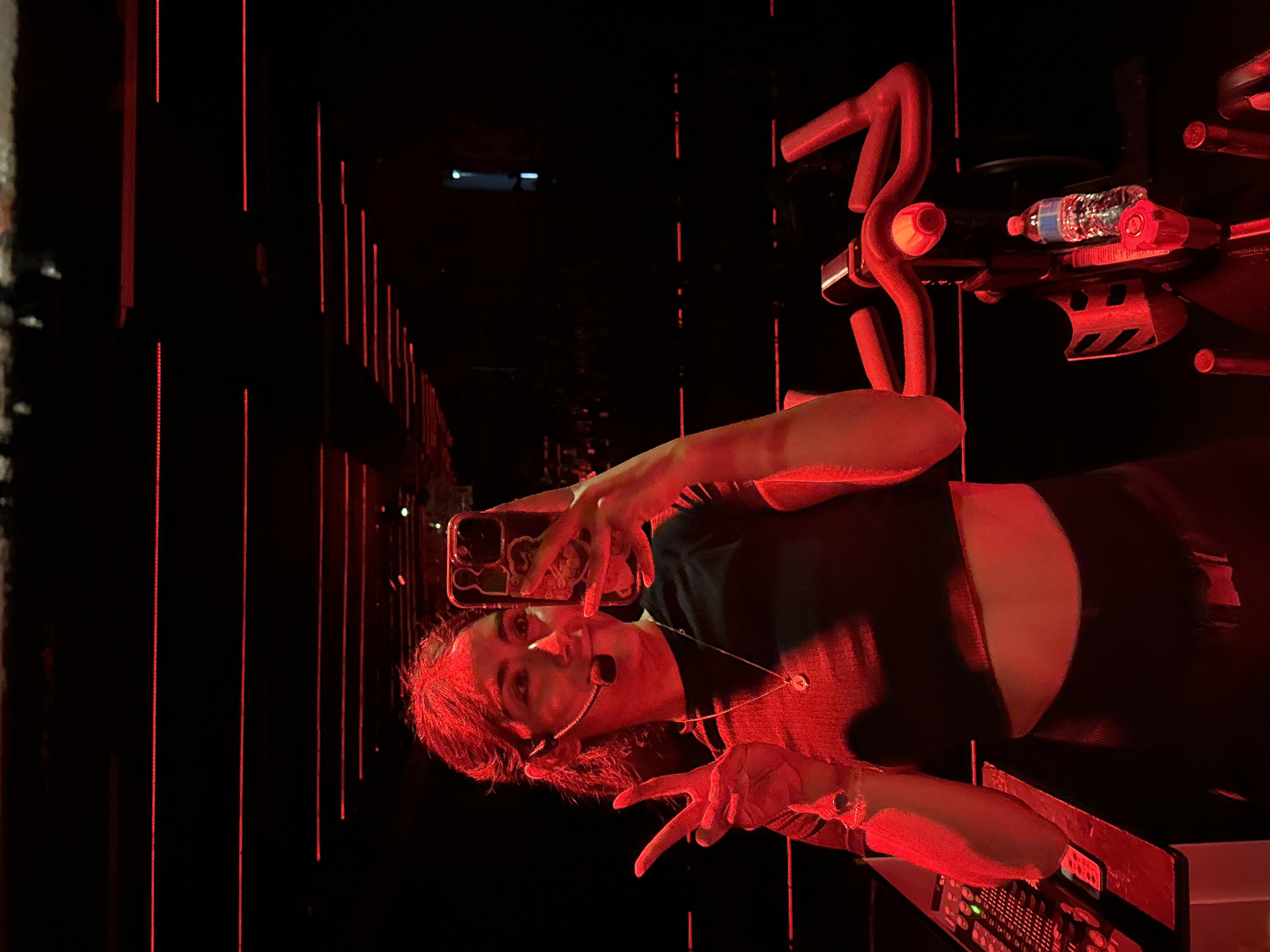 A woman is taking a selfie with her smartphone next to a stationary exercise bike in a dark room with red vertical lines of light. She is wearing a black top and a headset microphone, and making a peace sign with her right hand. The exercise bike has a water bottle on it. The room appears to be a fitness studio. It is impossible to determine the location of the city of Puebla, Mexico, from the image.