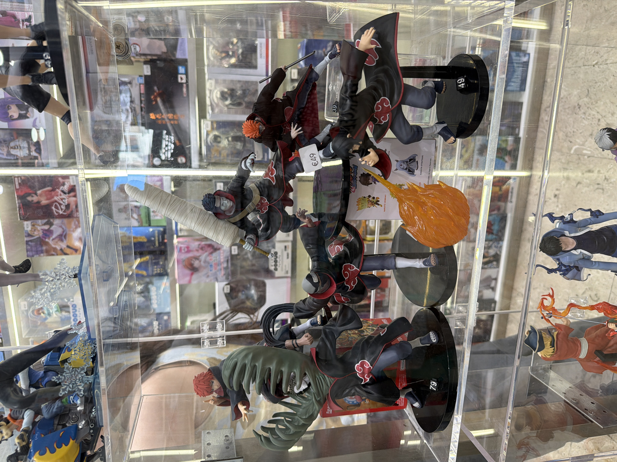 The image displays a collection of anime action figures displayed in a glass-enclosed shelf, likely in a retail environment. The figures are primarily from the anime series Naruto, characterized by their distinctive black cloaks with red cloud emblems. Several figures depict characters in dynamic poses, suggesting combat or action sequences. One figure, in the foreground to the left, has bright red spiky hair and wears a green, tentacle-like accessory. Another figure, slightly behind and to the right, is positioned as if striking with a weapon. In the middle ground, a figure with orange hair and purple facial markings is shown holding a staff. A price tag reading "€49" is visible near this figure. Below the main grouping, more figures are visible, including one with an orange flame effect erupting from its hand and another with a black and grey hooded outfit. The background reveals rows of other anime merchandise and displays. The setting appears to be a shop or a collector's display. There are no people directly visible interacting with the figures. Based on the presence of the Milanese fashion brand "Prada" and a price tag using the Euro currency, it is highly probable that this image was captured in Milan, Italy.