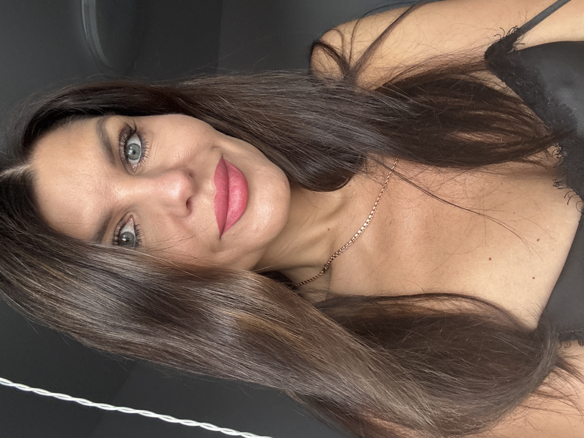 A woman with long, dark brown hair and blue eyes is pictured in a close-up, upper body shot. She is wearing a necklace and a black garment, likely lingerie. The background is dark and nondescript, with a white, twisted cord visible on the left side of the frame. The lighting is soft, highlighting her facial features and the texture of her hair and skin. There is no discernible urban or natural environment visible, nor any indication of the city of Chaikovsky, Russia.