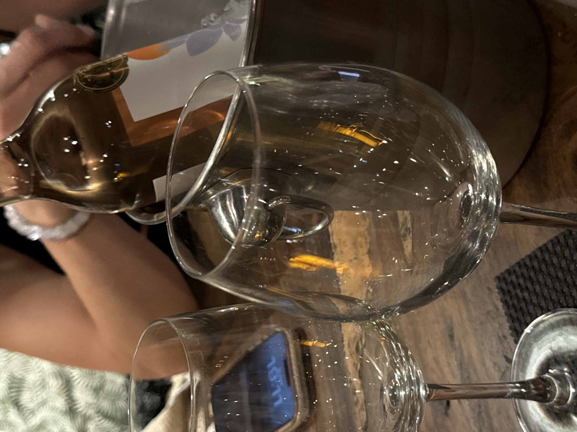 A close-up shot captures two wine glasses, one partially filled, alongside a bottle of wine, likely on a wooden table. The image is likely taken in Ubud District, Indonesia. The scene suggests a dining or social setting, potentially in a restaurant or private residence. A person's arm and wrist are visible in the periphery, possibly holding the bottle, indicating interaction with the wine.