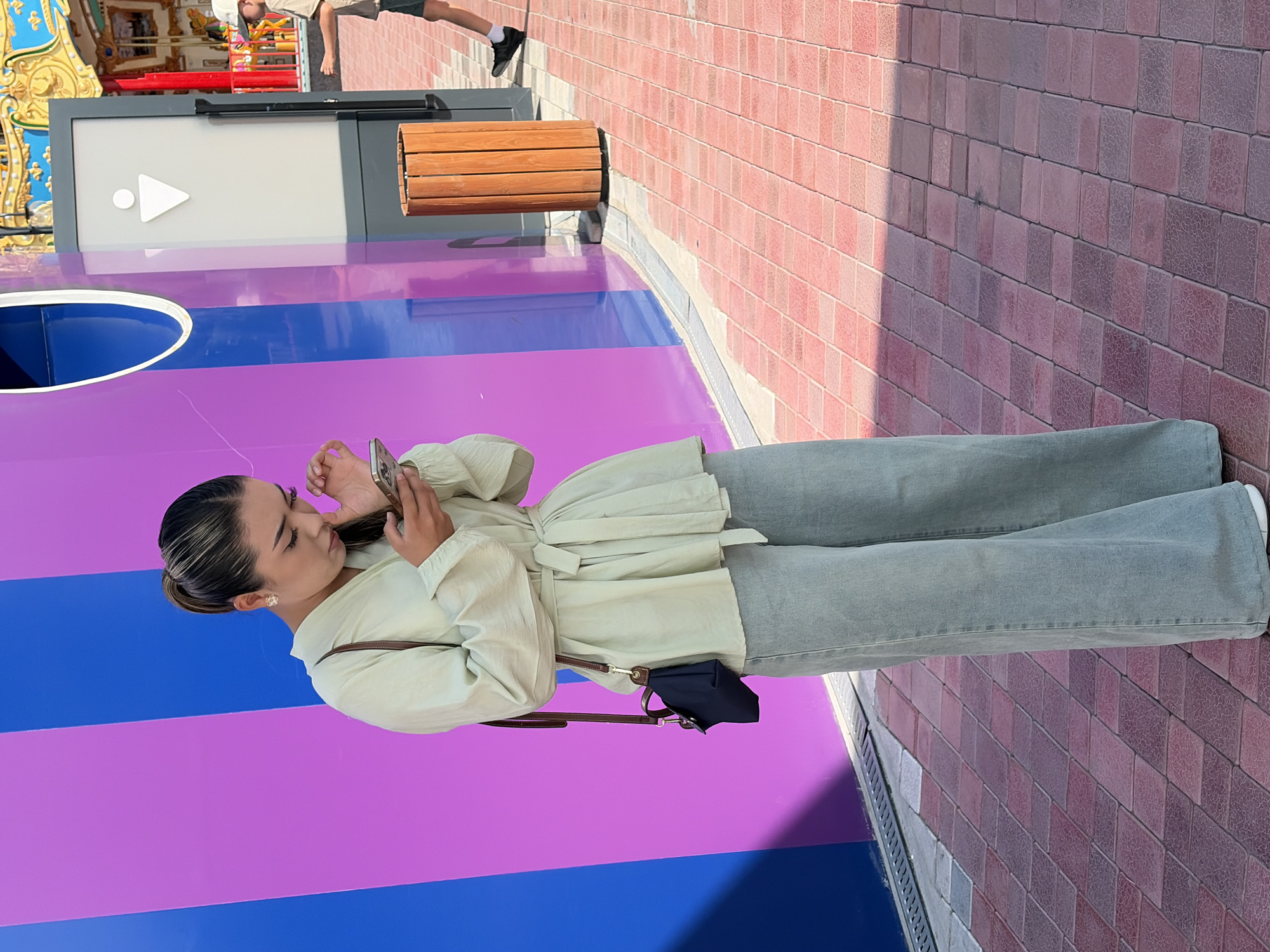 A full-body, slightly angled shot shows a young woman with her hair pulled back in a ponytail, standing outside and looking at her smartphone. She is wearing a light green, long-sleeved, collared shirt with a tie belt at the waist and light blue wide-leg pants. A dark blue crossbody bag with a brown strap hangs from her left shoulder. She is standing on a brick-paved walkway in front of a wall with vertical stripes of blue and purple. To her right, there is a cylindrical wooden trash can and a structure with a male restroom symbol on it. A carousel is visible in the background. The lighting suggests it is daytime.