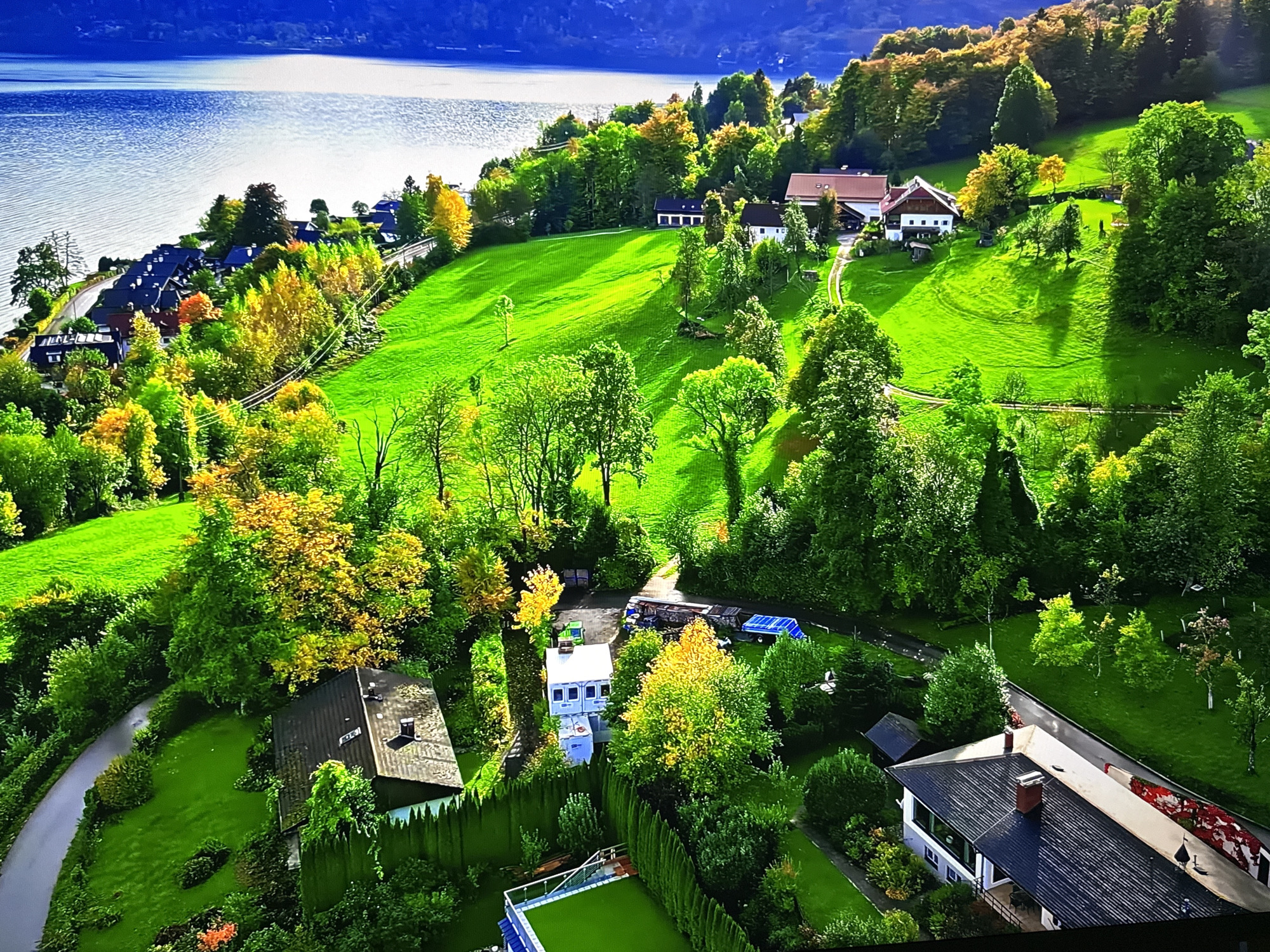 This aerial drone footage captures a picturesque, verdant landscape bordering a large body of shimmering blue water, possibly a lake or sea. The scene is characterized by rolling hills covered in lush green grass, interspersed with dense patches of trees exhibiting vibrant autumn foliage in shades of yellow, orange, and green. Several modern residential buildings with dark-pitched roofs are nestled into the landscape, some appearing to be clustered together along a winding road that leads down towards the water. One prominent building features a white facade and dark roof, with visible chimneys. Further inland, other structures are visible, including smaller outbuildings and what appears to be a fenced sports court with artificial turf. A grey asphalt road snakes through the scene, connecting different areas and providing access to the houses. The overall impression is one of a serene, affluent rural or semi-rural setting, likely a residential area in a scenic location. However, there is no visual information within the image to suggest this location is the city of Burgas, Bulgaria.