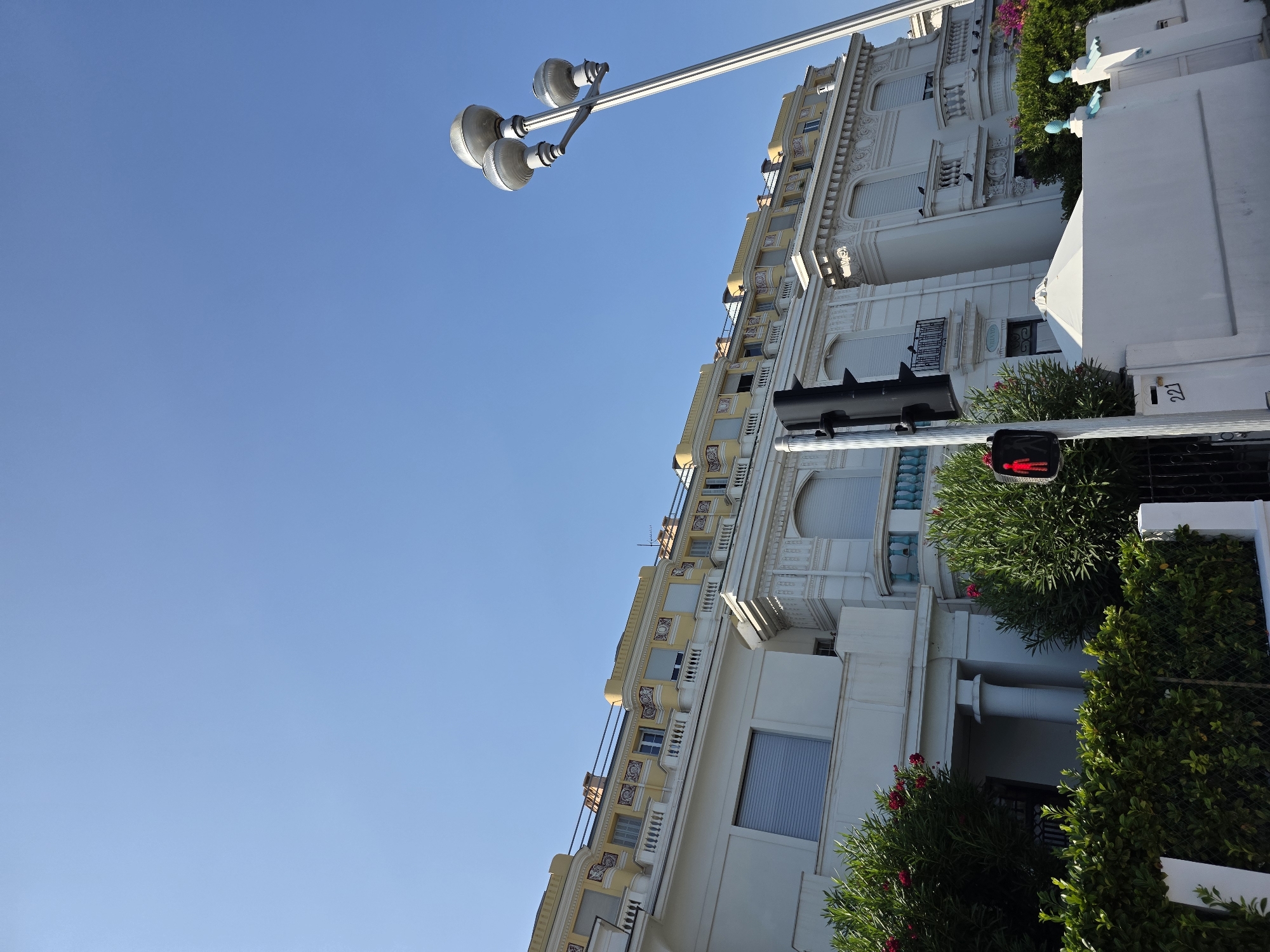The image captures a view of a multi-story building with a yellow facade on the upper floors, characteristic of the architecture found in Nice, France. The building features ornate detailing, balconies with white railings, and numerous windows with closed shutters. In the foreground, a traffic light displays a red pedestrian signal, indicating "don't walk." Green hedges and trees, some with red flowers, line the front of the property, partially obscuring the lower levels of the building. A white fence and a lamppost with three globes are also visible. The sky is clear and blue. The scene appears to be a street view in a sunny urban environment.