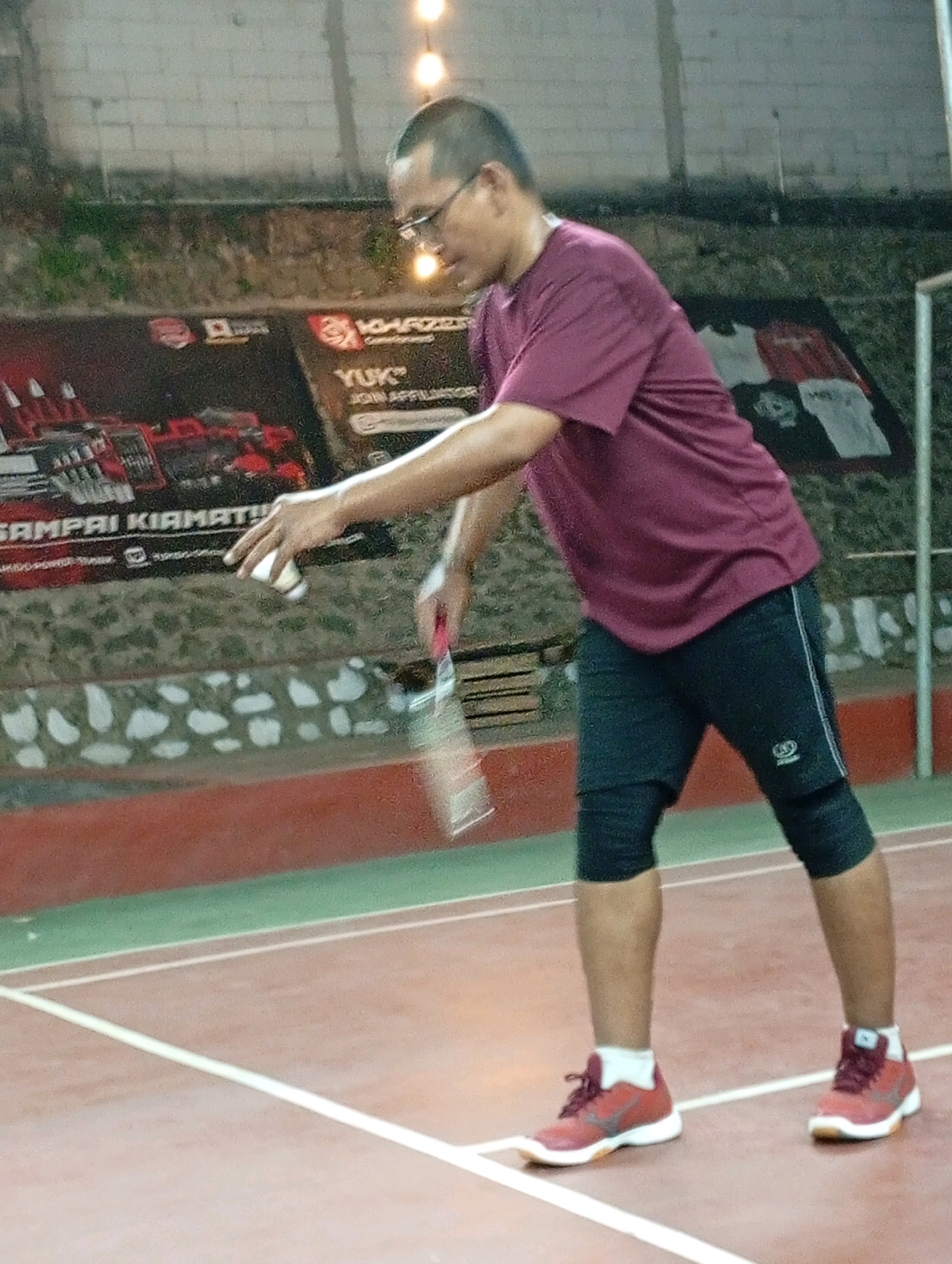 An adult male with short hair and glasses is playing badminton indoors. He is wearing a maroon t-shirt, black shorts, black knee pads, and red athletic shoes. He is in the motion of hitting a shuttlecock with a badminton racket. The background shows a brick wall with posters and lights. The court floor is red with white lines. The image appears to have been taken at a badminton court in Caringin, Indonesia.