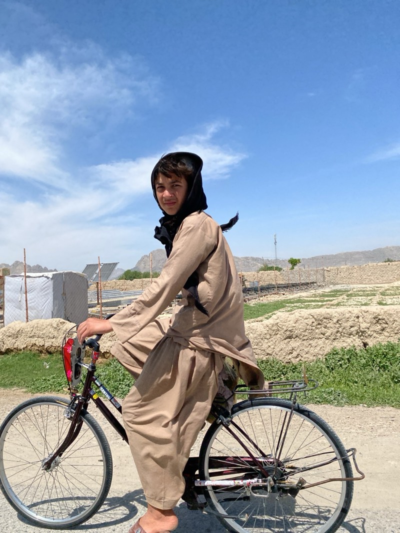 A young person rides a bicycle in what appears to be an outdoor, arid environment. The individual wears a light brown traditional outfit, and a dark covering on their head. The bicycle is an older model. The background shows a landscape with a settlement, possible agricultural areas, and a dry, hilly terrain. The sky is blue with scattered clouds. The location is consistent with the general environment and architecture found in regions like Kandahar, Afghanistan.