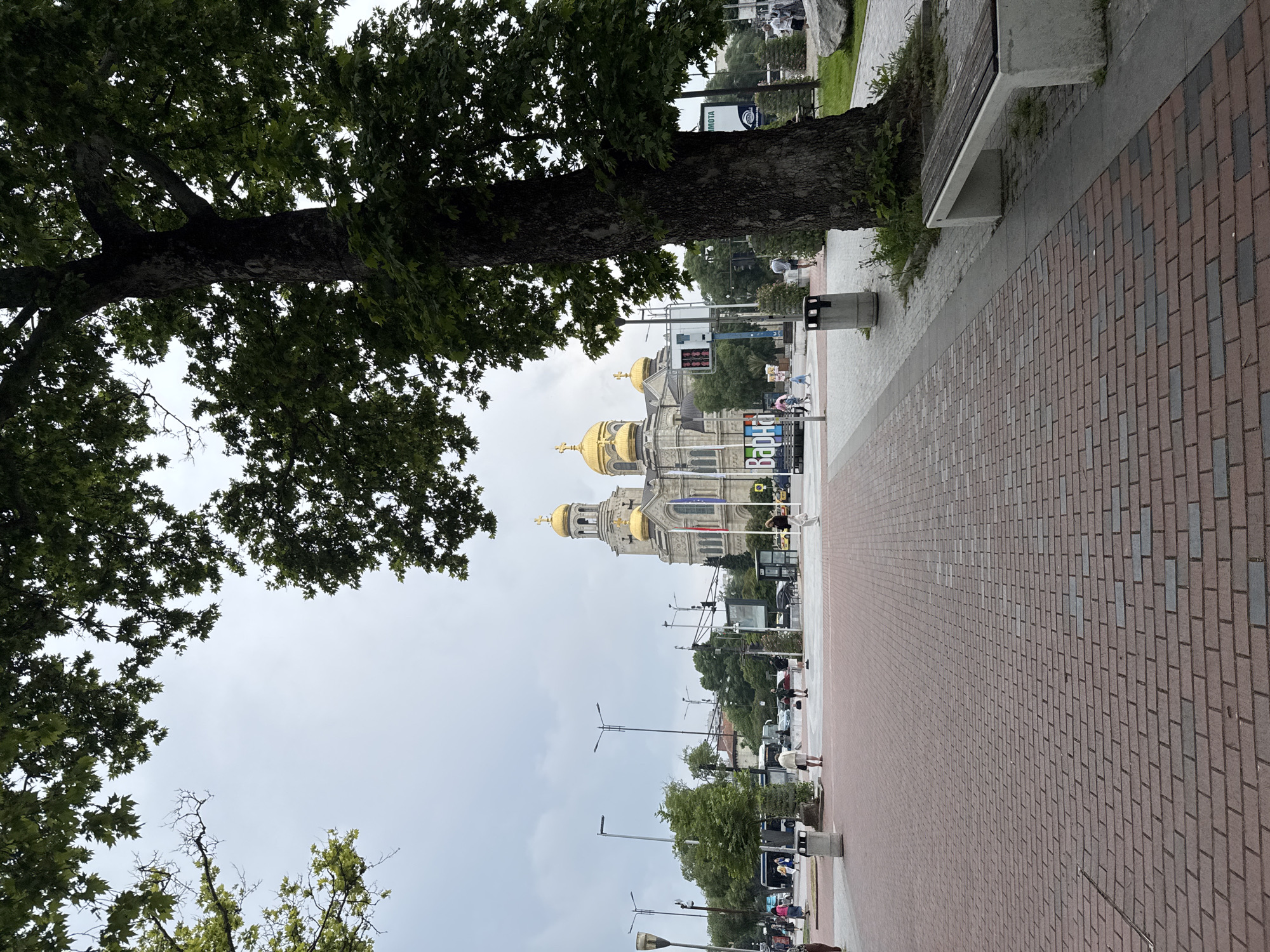 This media file captures an urban scene featuring a prominent cathedral, a large tree, and a pedestrian pathway on an overcast day.
In the foreground and left, a large, mature tree with a thick, textured trunk and dense green foliage dominates the left side of the frame, extending its branches upwards and across the top. The leaves are broad and deeply lobed.
To the right of the tree, a wide pedestrian pathway stretches into the background. It is paved with red and grey brick-like tiles laid in a herringbone pattern. Along this pathway, several modern amenities are visible, including concrete benches with wooden slats and dark grey cylindrical trash cans.
In the mid-ground, a grand, ornate building with light-colored stone architecture and multiple striking golden domes, topped with crosses, stands as a central feature. This building appears to be an Orthodox cathedral. Several flags are displayed on its facade.
Beyond the pathway and around the cathedral, various people can be seen walking or standing, some with bicycles. Streetlights and other urban infrastructure, like traffic lights and signs, are also present. Vehicles are partially visible in the background, suggesting a street or road runs alongside the pedestrian area.
The sky is uniformly grey and overcast, indicating a cloudy daytime. Visible text on signs includes "Варна" (Varna) and "МОТА" (Mota), providing a strong clue about the location. The overall impression is a bustling public square or promenade in a European city, with the cathedral serving as a significant landmark.