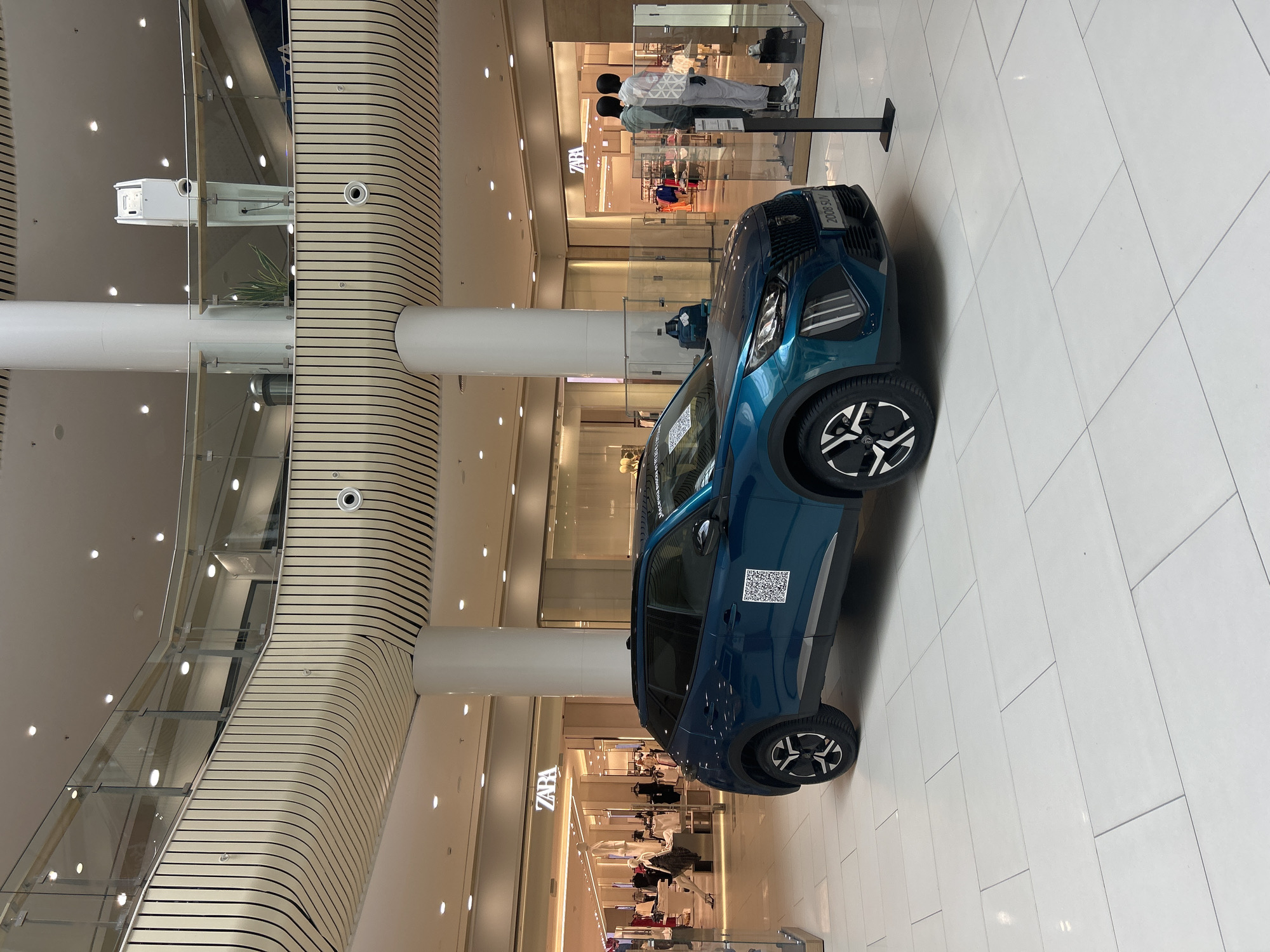 The image shows a blue 2008 SUV car placed indoors on a white tiled floor in what appears to be a shopping mall in Plovdiv, Bulgaria. The car's front is visible, with the words "2008 SUV" printed on the front. A QR code is visible on the car's side. In the background, there are storefronts of retail stores, with Zara being one of them, featuring mannequins and clothes. The mall structure includes vertical wooden-striped support beams and glass railings for upper levels. The lighting is bright and even, suggesting artificial lighting within the mall.