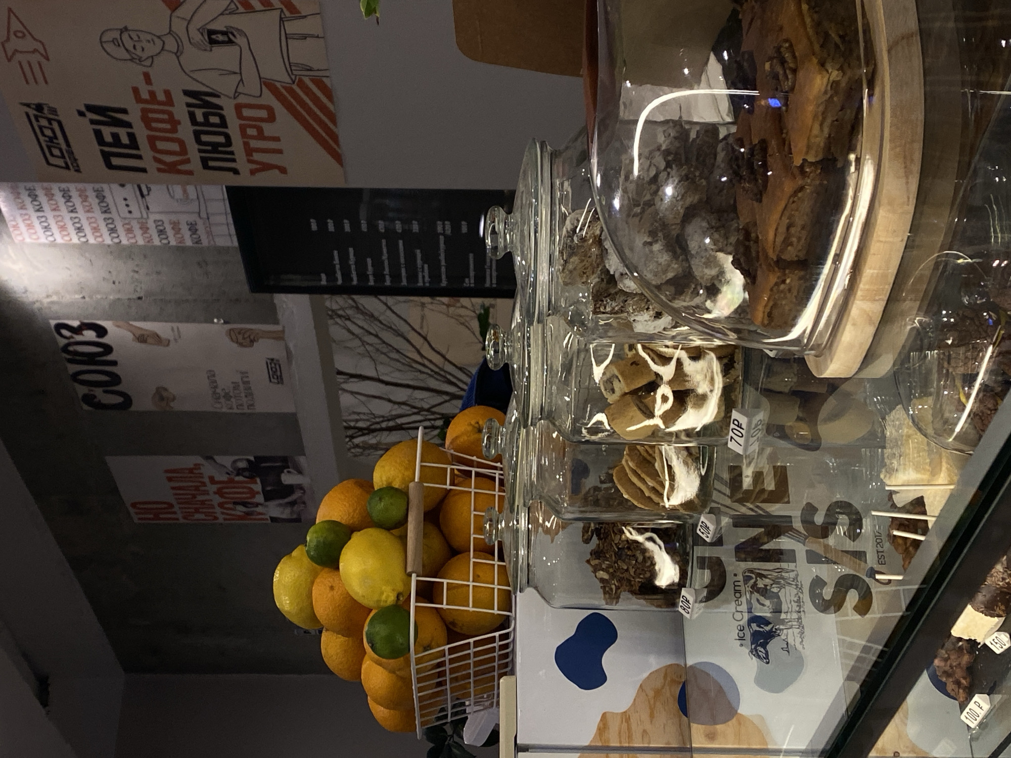 The image depicts an interior scene, likely a cafe or a similar establishment, with a focus on food and beverages. In the foreground, a glass display case showcases various food items. To the left, a selection of fruits, including oranges, lemons, and limes, are piled in a white basket. Behind the fruit, there are several large glass jars filled with what appears to be granola or cereal. Further to the right, a glass dome covers a tray of baklava, a pastry dish. Reflections on the glass surfaces reveal the surrounding environment, including what appears to be a menu board with prices. On the wall in the background, there are posters with Cyrillic text and graphics. One poster features a stylized rocket and the text "СОЮЗ", another has text that translates to "Drink Coffee - Love Morning". The overall impression is that of a food service establishment, possibly a cafe or bakery, with a modern aesthetic. The provided context identifies the location as Vishnyaki, Russia, suggesting this is likely a local business.
