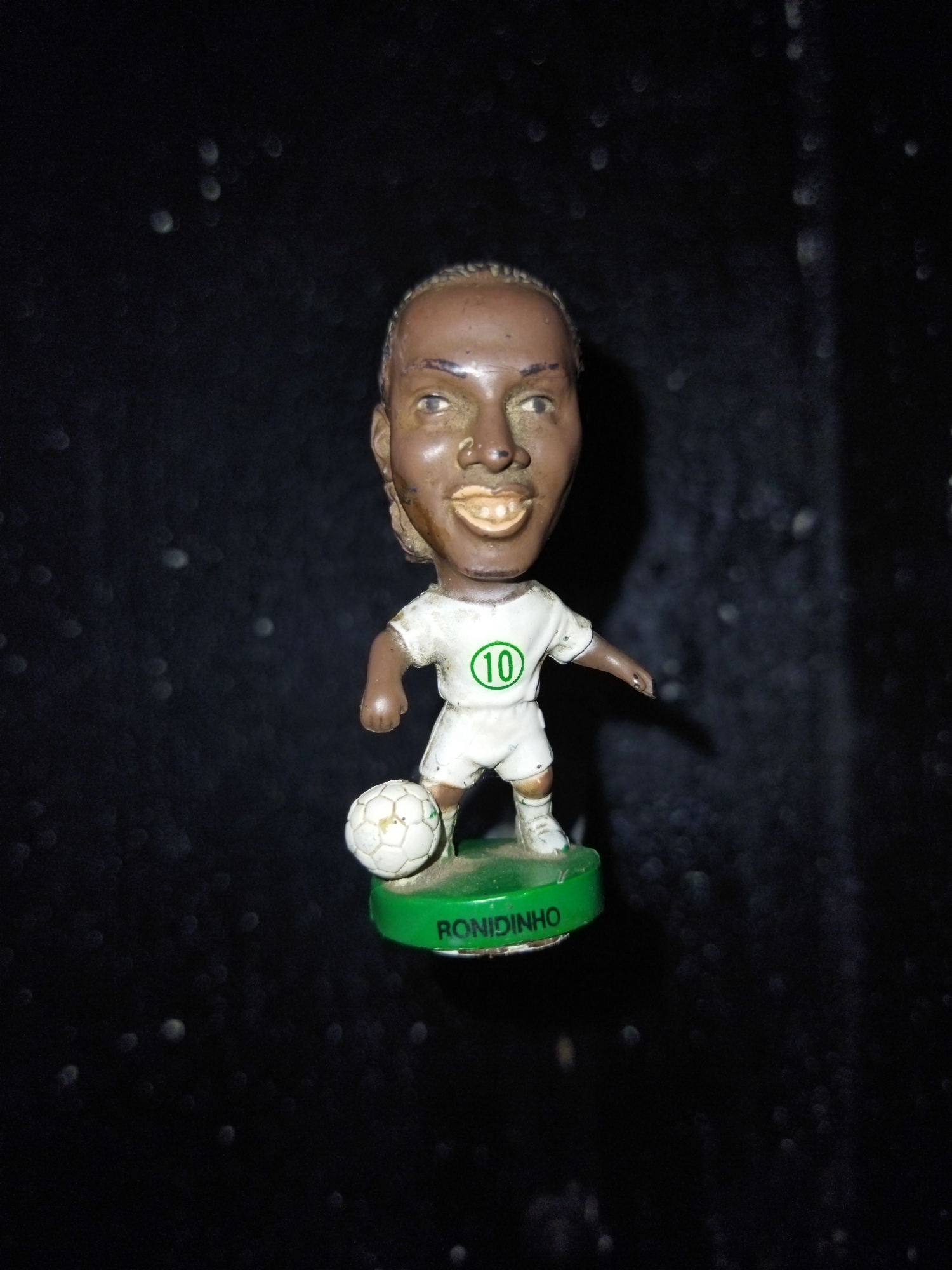 The image shows a small, painted figurine of the soccer player Ronaldinho, wearing a white uniform with the number "10" in a green circle, standing on a green base with his name printed on it. He is next to a soccer ball. The background is black. The image does not provide information about location in the city of Tinaquillo, Venezuela.