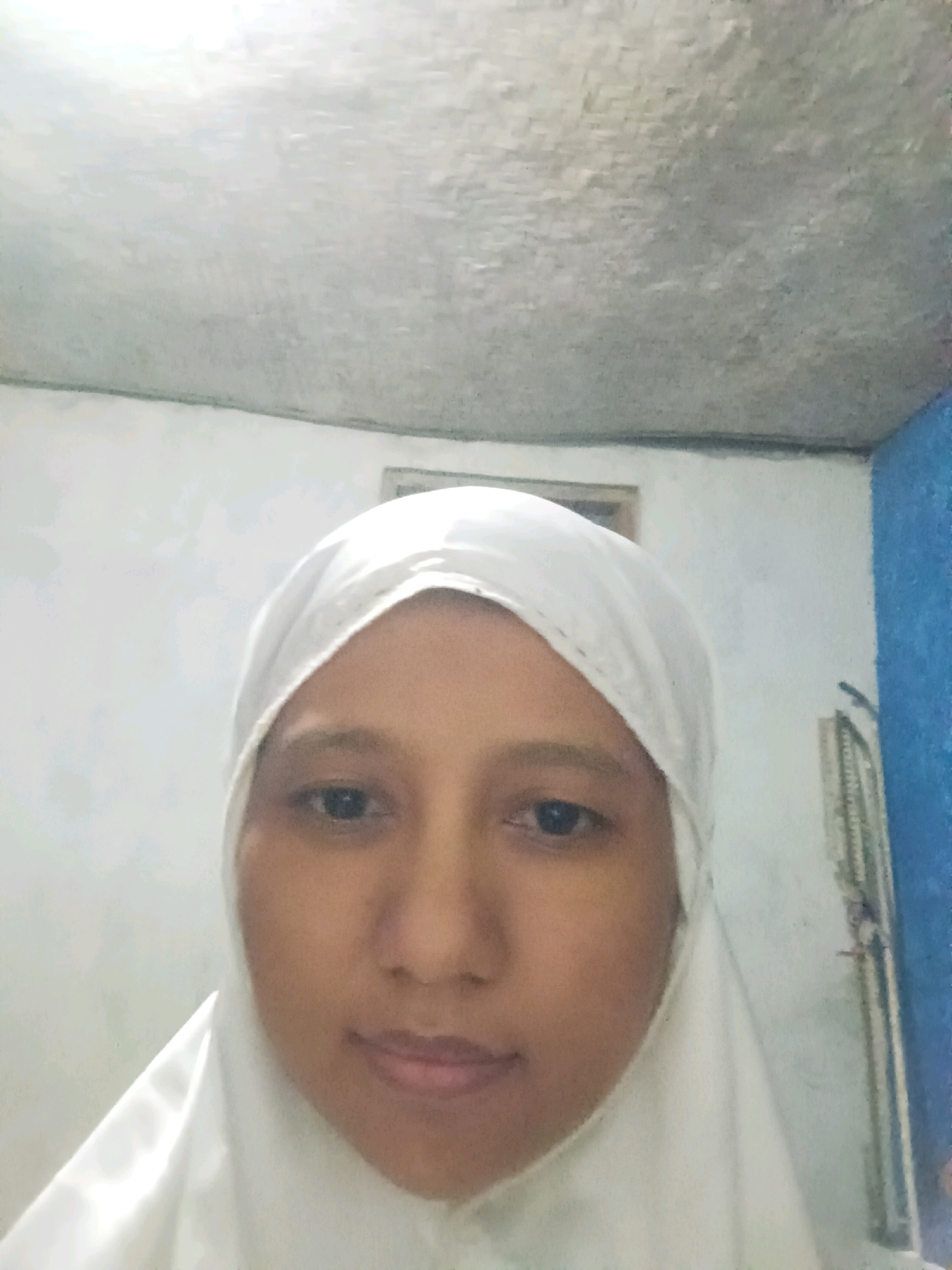 A medium shot selfie is captured from a slightly low angle of a young woman wearing a white hijab. She is looking directly at the camera with a neutral expression. The background consists of a textured white wall and ceiling, with a blue-painted wall and a vertical structure, possibly a door frame or a cabinet, visible on the right side of the image. The lighting is somewhat diffused, casting a soft glow on the subject's face. The scene appears to be indoors, likely in a residential setting in Canga'an, Indonesia.