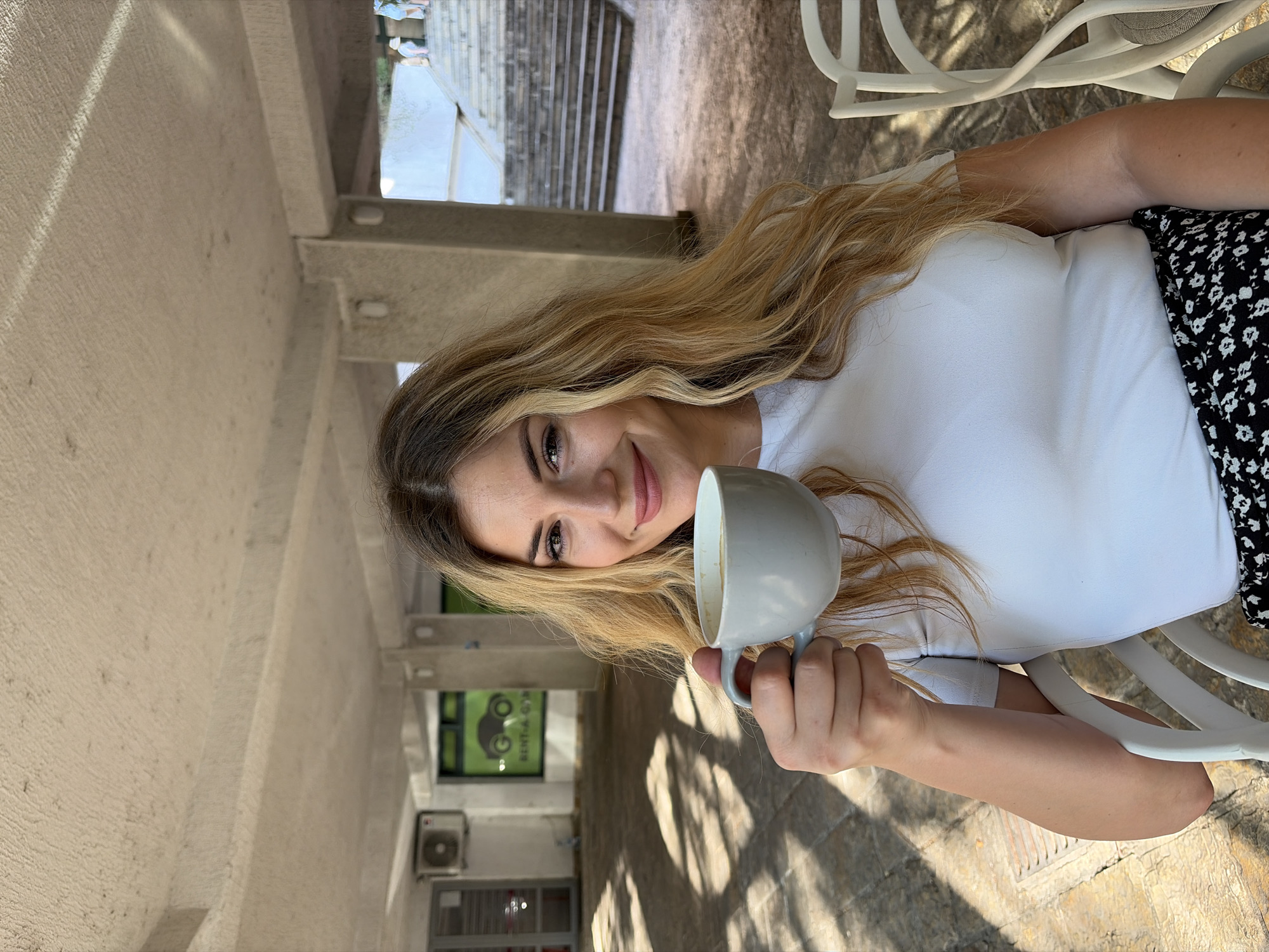 A young woman with blonde, wavy hair sits at an outdoor cafe in Budva, Montenegro, holding a light grey mug of coffee. She is wearing a white t-shirt and a black and white patterned skirt. The cafe appears to be situated under a covered walkway with a textured ceiling. In the background, a building with an air conditioning unit is visible, along with a window displaying a "RENT-A-CAR" sign with a car graphic. Further in the background, a wide set of outdoor stairs leads up to a higher level. The ground is paved with textured stones, and sunlight casts shadows across the scene. White plastic chairs are visible in the foreground and to the right.