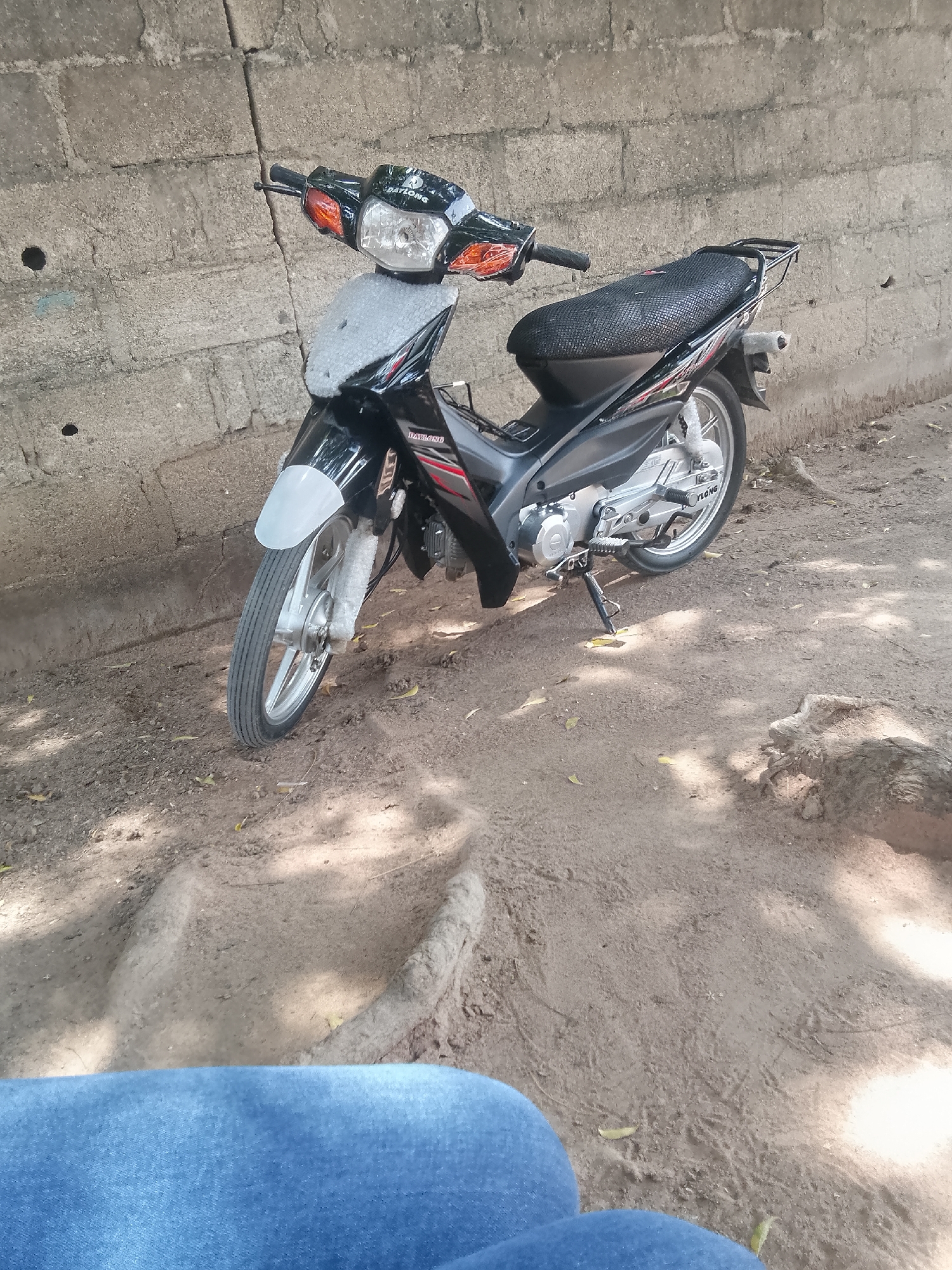 A black motorcycle with a silver front fender and silver wheels is parked on a dirt path in front of a textured concrete block wall. The motorcycle has a black seat with a textured cover and a rear rack. The front of the motorcycle is partially covered with a light-colored material, possibly protective wrapping. The ground is a mix of dirt and small stones, with some dry leaves scattered around. Sunlight casts dappled shadows on the ground. The scene appears to be outdoors, likely in a residential or storage area. The image has been taken from a slightly elevated position, looking down at the motorcycle and the ground. The lower portion of the image shows the blue denim of someone's jeans, suggesting the photographer is sitting or kneeling. The overall impression is one of a still, everyday scene. The presence of the motorcycle and the style of the wall suggests a location in Jalingo, Nigeria, where such vehicles are common for transportation and the building materials are typical.