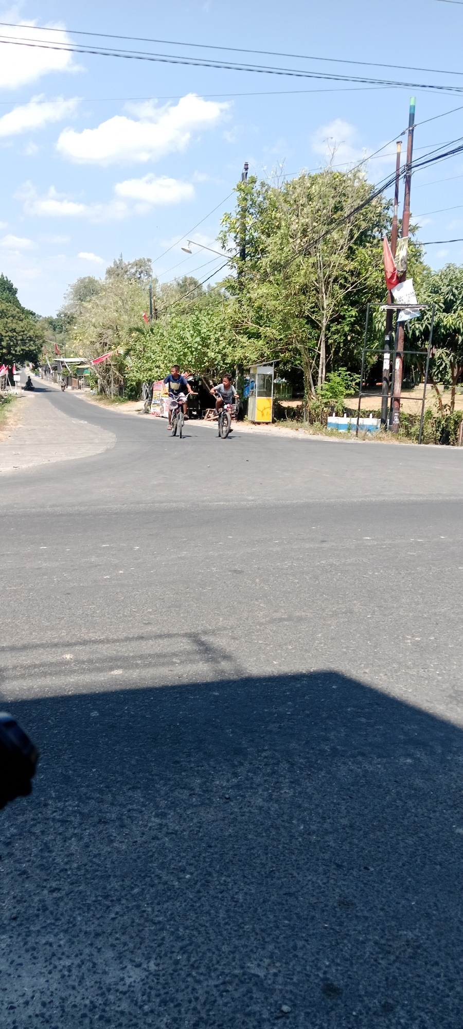 Two young boys are riding bicycles on an asphalt road under a clear blue sky with scattered clouds. The boy on the left is wearing a dark blue t-shirt and white shorts, while the boy on the right is wearing a grey t-shirt and dark shorts. They are riding side-by-side, approaching a slight bend in the road. The road is surrounded by lush green trees and vegetation. To the right of the road, there is a yellow structure, possibly a small kiosk or booth, and utility poles with numerous wires. In the background, there are buildings with red roofs and more trees. The scene appears to be in a semi-rural or suburban area, consistent with the location of Maguwoharjo, Indonesia, which is known for its mix of residential areas, natural landscapes, and some commercial establishments. The lighting suggests it is daytime, likely late morning or early afternoon. The overall atmosphere is peaceful and ordinary, depicting children enjoying outdoor activity.