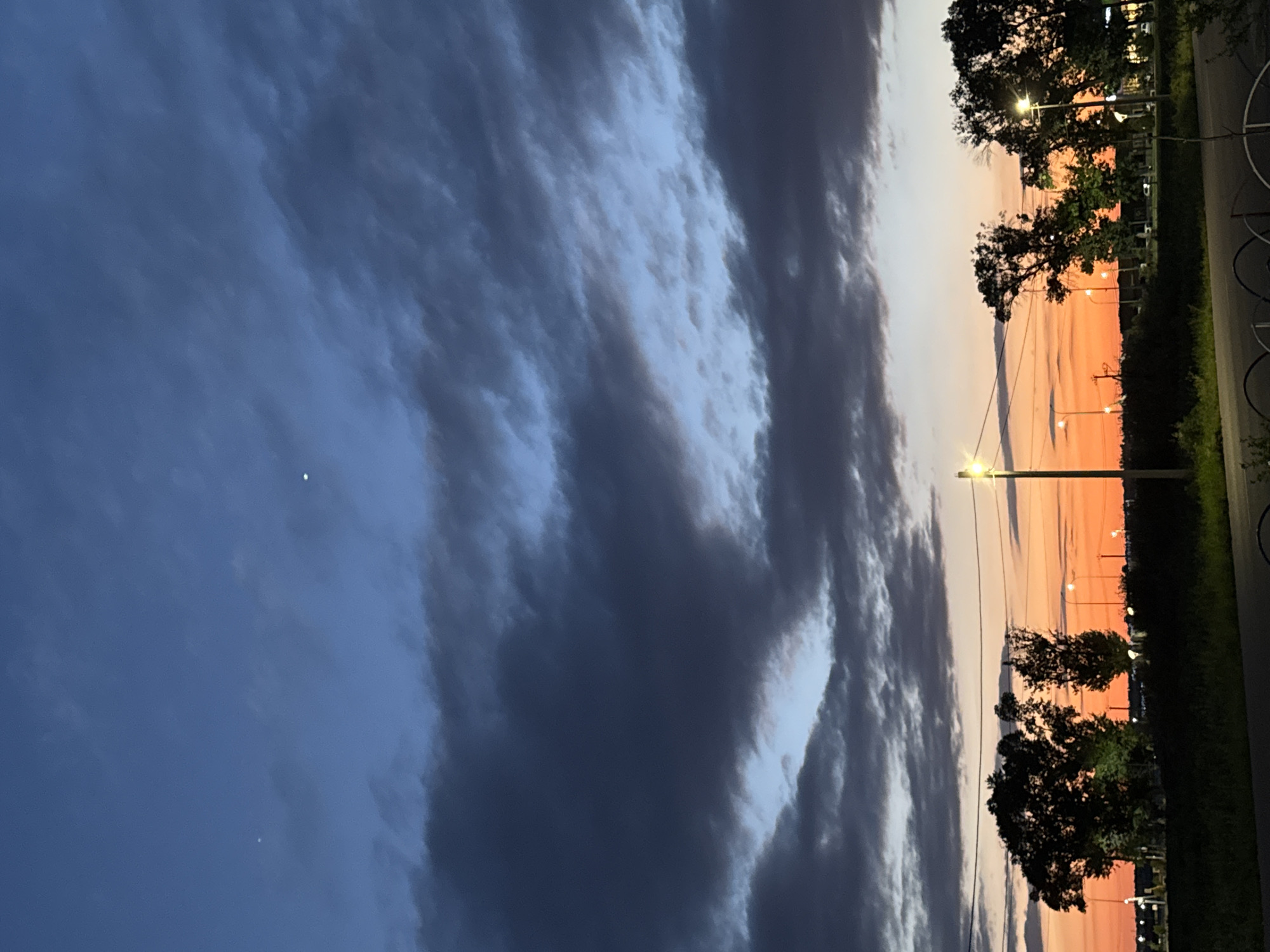 The image shows a twilight sky over a suburban or exurban landscape. The sky is a gradient of deep blue at the top transitioning to a vibrant orange and pink near the horizon. Dark, dramatic clouds are scattered across the upper portion of the sky. In the foreground, silhouetted trees and vegetation line a slightly elevated grassy area. A road or pathway is visible in the very bottom of the frame, with a dark metal fence or railing. Streetlights are illuminated along the horizon, suggesting the presence of a road or parking lot in the distance. Two small, bright celestial bodies are visible in the upper part of the sky. The scene appears to be devoid of people or vehicles.