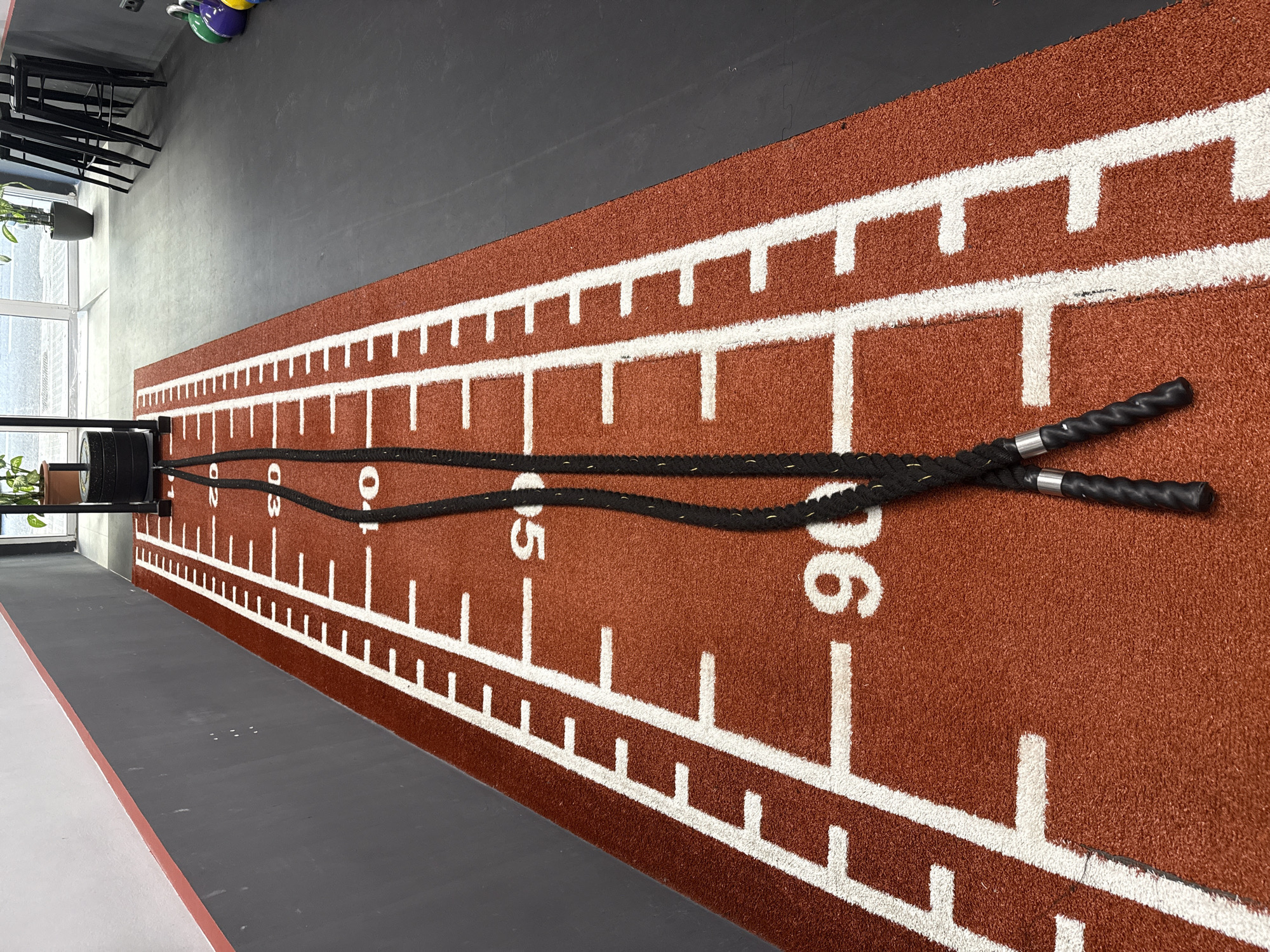 The image captures a modern gym or fitness studio floor, featuring a prominent reddish-brown athletic track marked with white lines and numbers. A black battle rope, with textured black handles and silver bands at its ends, lies doubled over on this track, extending along its length. The rope appears anchored to a black metal frame or weight rack, partially visible at the left end of the track.
The track itself is marked with white horizontal and vertical lines, resembling a measuring tape, with numbers "01", "02", "03", "04", "05", and "06" clearly visible, likely indicating meters. Adjacent to the track on one side is a darker gray floor, and on the other, a lighter gray or white floor section.
In the background, natural light streams in from a large window on the left. Stacked black chairs or stools are visible in the upper left corner, next to a potted plant. Colorful kettlebells (green, blue, yellow, purple) are stored in the upper background, indicating a functional training area. The scene depicts a well-equipped, organized fitness space, with the battle rope suggesting strength and conditioning exercises. The time of day appears to be daytime due to the natural illumination. The location context for this gym is Barcelona, Spain.