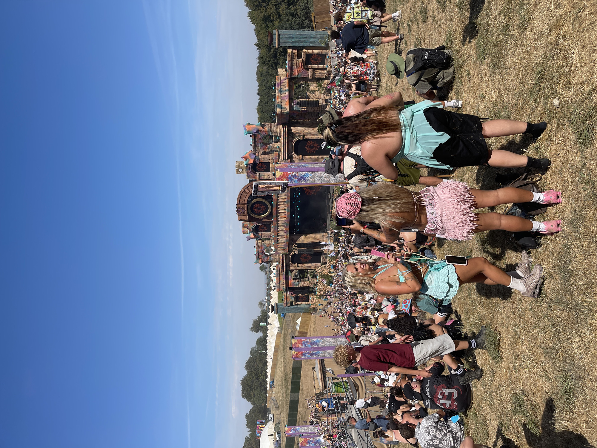 The image depicts a large outdoor music festival with a massive stage structure in the background, featuring ornate architectural elements and vibrant flags. A dense crowd of festival-goers is gathered, many sitting on the dry, sun-baked grass. In the foreground, several individuals are visible, some standing and others seated. The people are dressed in casual, summery attire, with some wearing festival-themed outfits. The sky is clear and blue, indicating a sunny day. The environment appears to be a field with some trees in the distance, and tents or temporary structures are visible on the far left. The overall scene suggests a lively and crowded event taking place during daylight hours. This location is the Matterley Bowl estate near Winchester, United Kingdom, which hosts the Boomtown Fair festival.