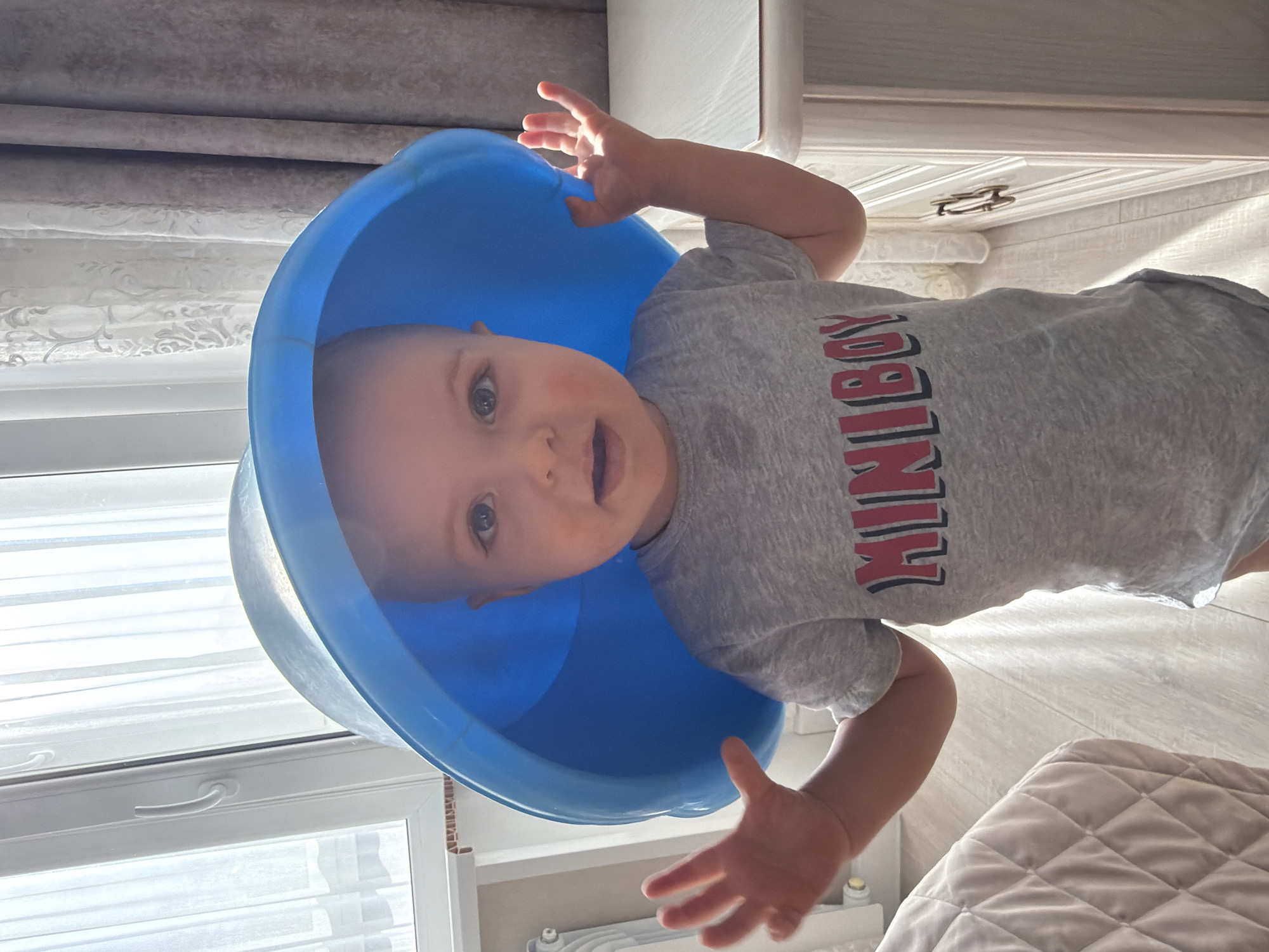 A baby wearing a gray t-shirt with the word "MINIBOY" in red letters has a blue plastic bucket over its head. The location is likely indoors, as suggested by the presence of a window, curtains, a radiator, a white cabinet, and a quilted bed cover. The baby is holding its hands up, seemingly attempting to remove the bucket. The image appears to be taken at an angle, possibly by someone looking down at the child. The setting is likely a residential area. The image contains no indication that it was taken in Votkinsk, Russia.