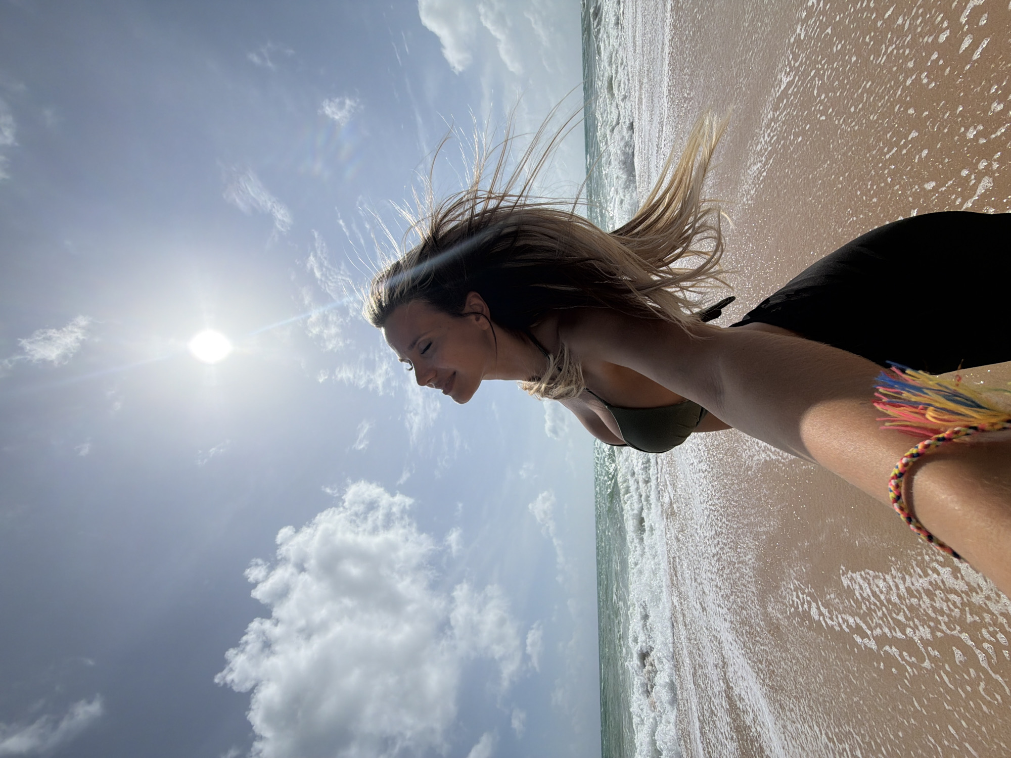 A young woman is standing on a beach in Mai Khao, Thailand. She is wearing a dark green bikini top and black bottoms. Her hair is blonde and blowing in the wind. She is looking up at the sun with her eyes closed. The ocean is behind her, with waves crashing on the shore. The sky is blue with some white clouds. A hand with a colorful bracelet is in the foreground, holding what appears to be a camera, taking a selfie.