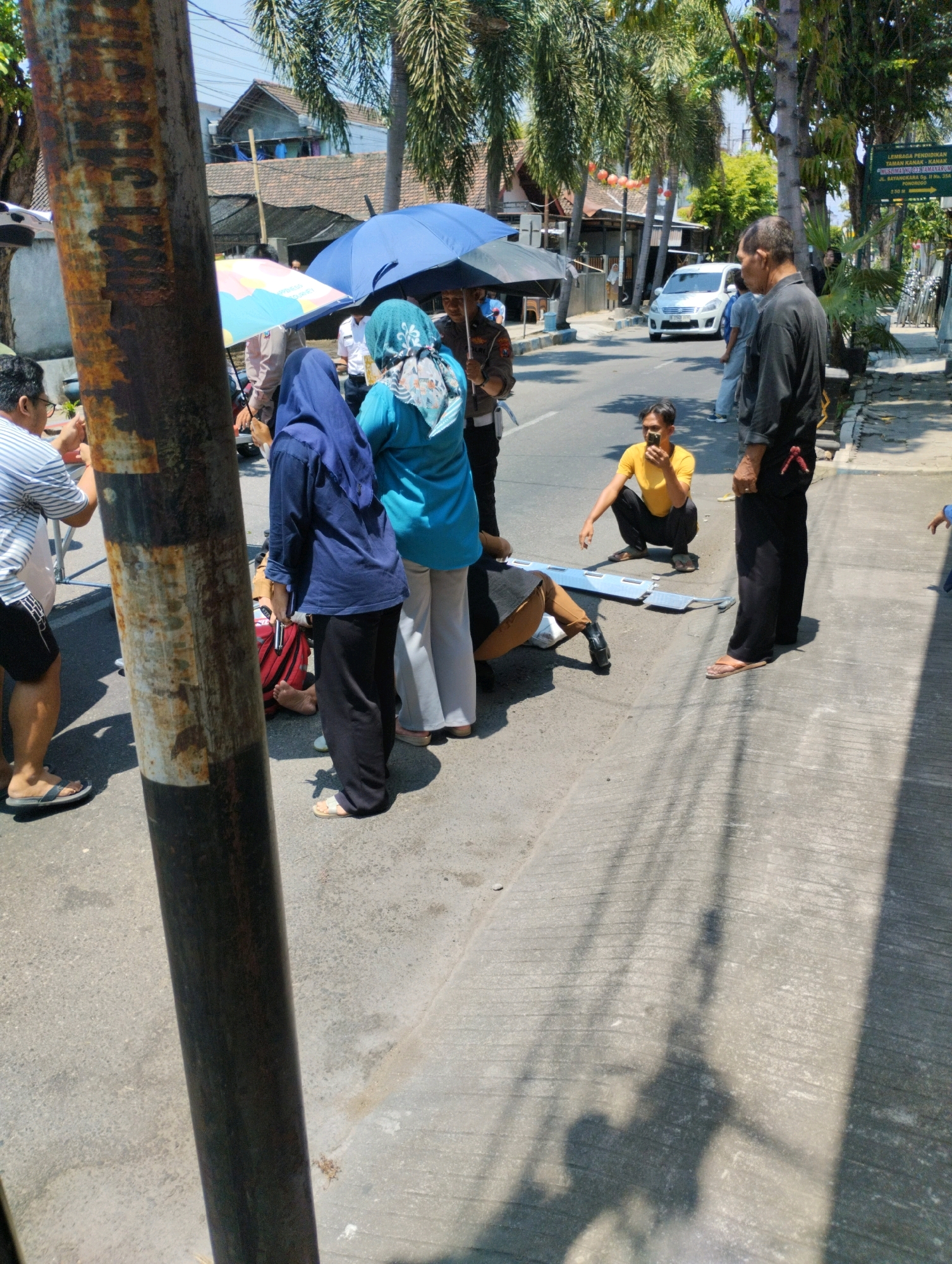 A group of people are gathered on the side of a street in Ponorogo, Indonesia, near a building with a terracotta roof and palm trees. In the foreground, a rusty pole obscures part of the scene. A person in a blue hijab and dark pants stands near a person in a blue top and light-colored pants. Another person in a blue hijab is also present. A man in a black shirt and pants stands to the right, looking away from the camera. In the middle ground, a person in a yellow shirt crouches down, holding a mobile phone. A white car is visible on the street in the background. Several umbrellas are open, suggesting it might be sunny or drizzly. A stretcher-like object lies on the ground. The overall scene appears to be one of concern or a minor incident.